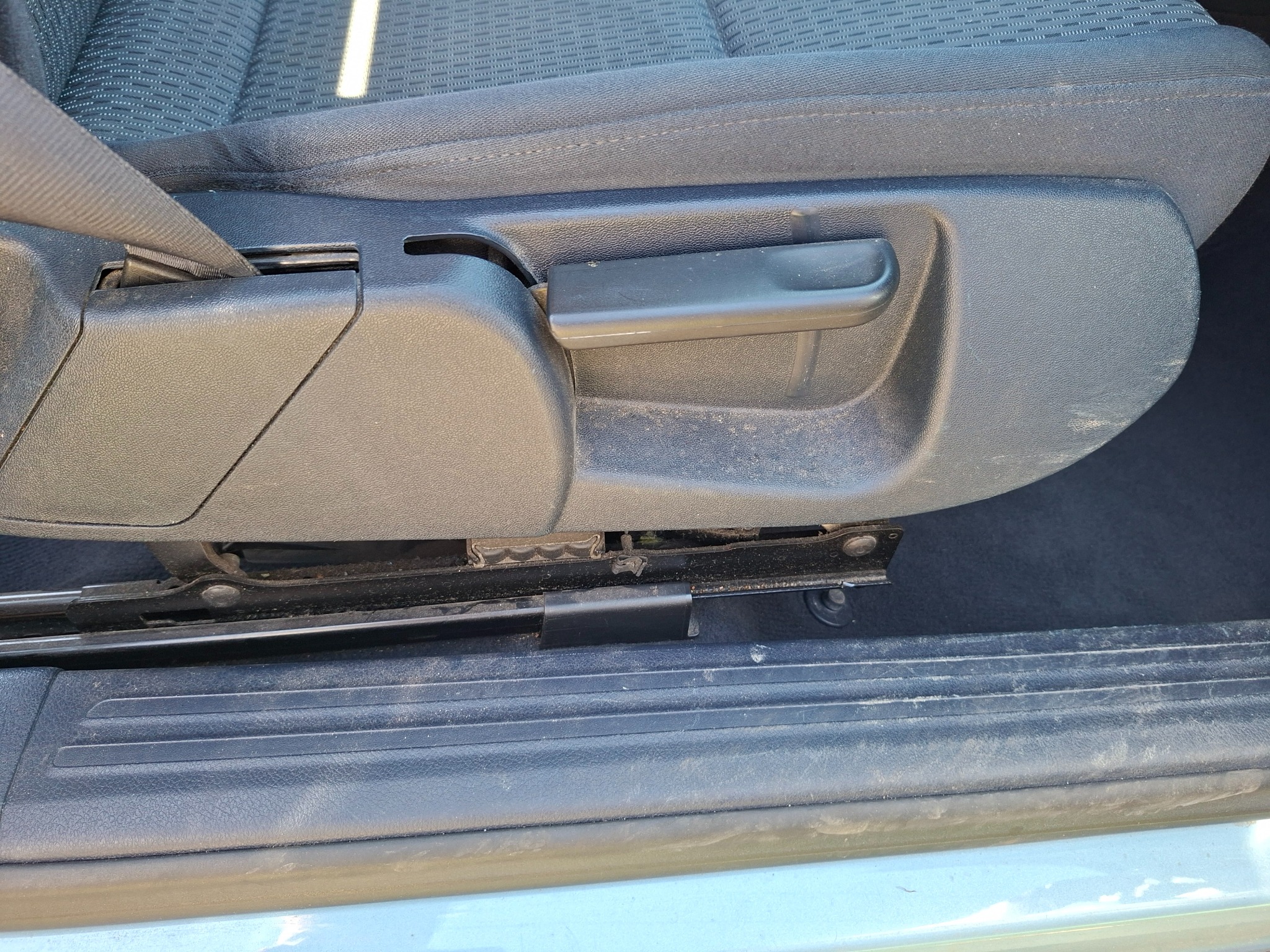 Brudne, tekstylne siedzenie samochodowe z pasem bezpieczeństwa i mechanizmem regulacji, widoczne zabrudzenia na tapicerce i elementach plastikowych.