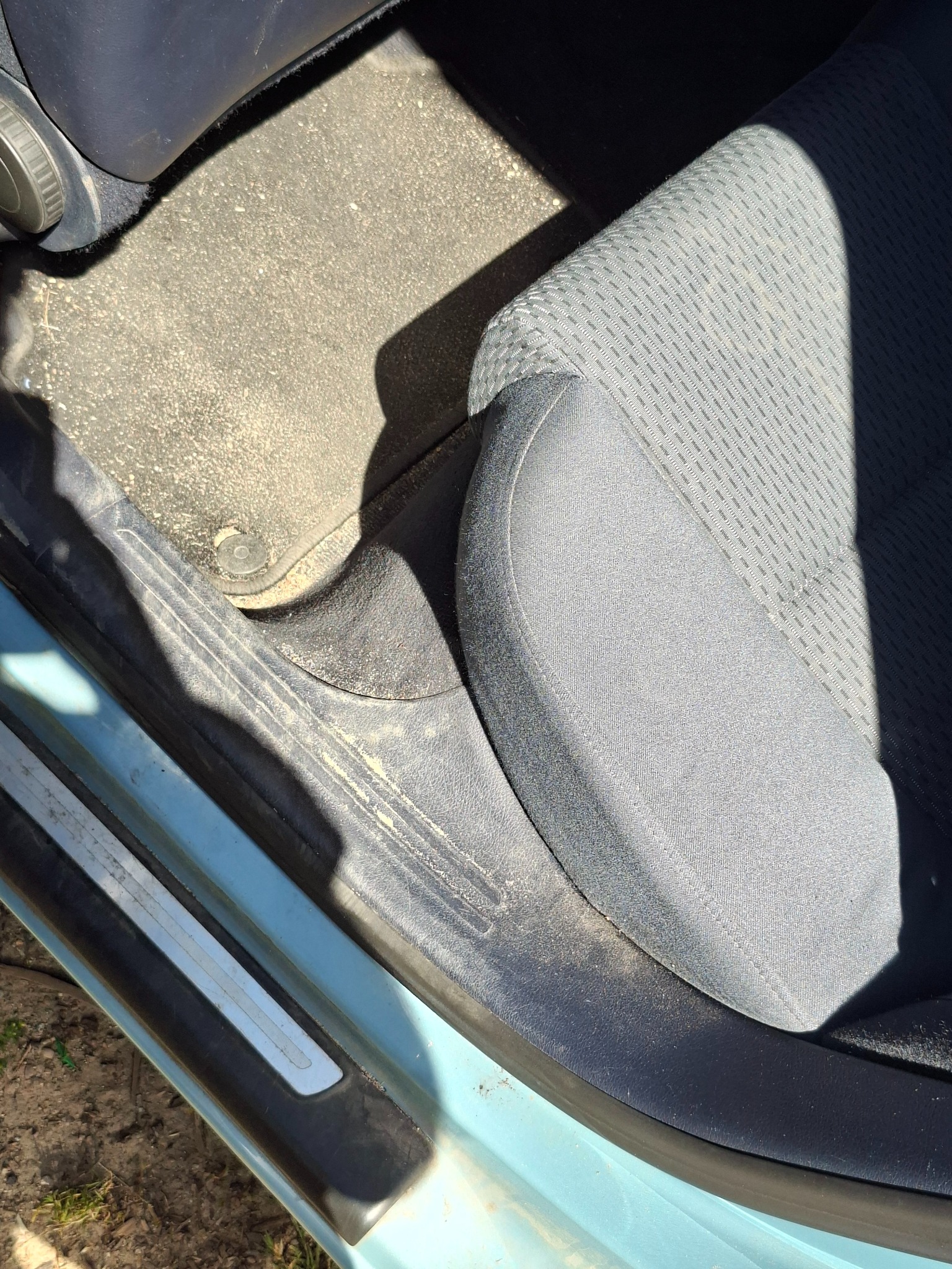 Wnętrze samochodu przed czyszczeniem: brudny dywanik, próg i fragment fotela. Widoczne zabrudzenia i kurz. Jasne, naturalne oświetlenie.