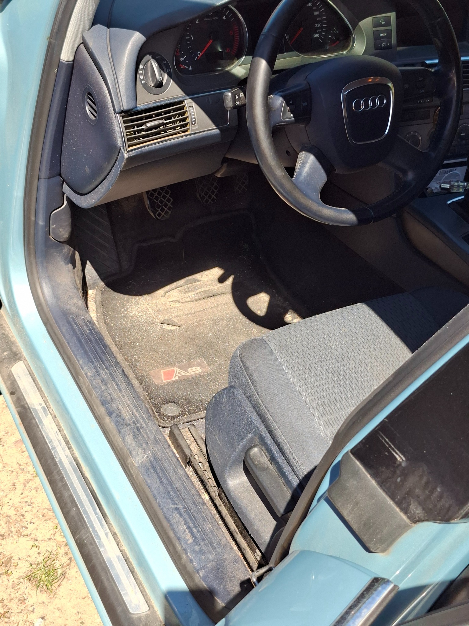 Wnętrze brudnego samochodu Audi A6, widok na fotel kierowcy, deskę rozdzielczą i dywaniki przed czyszczeniem. Widoczne zabrudzenia na elementach tapicerki.