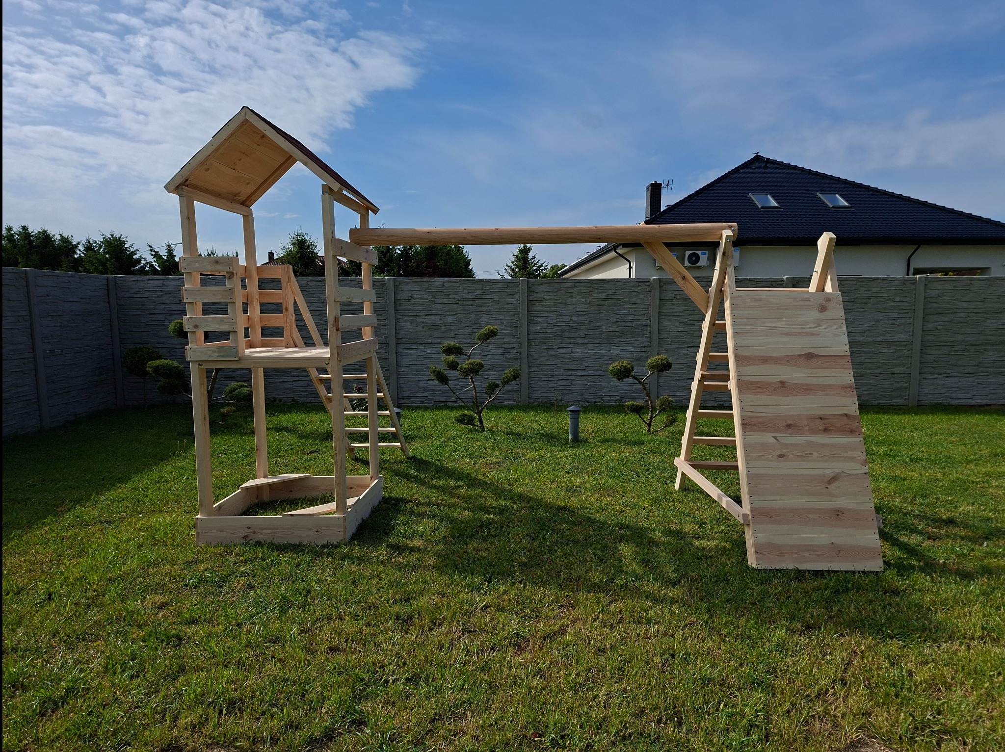 Drewniany plac zabaw z domkiem, zjeżdżalnią i drabinkami, ustawiony na trawniku przed betonowym ogrodzeniem. W tle dom z ciemnym dachem i błękitne niebo.