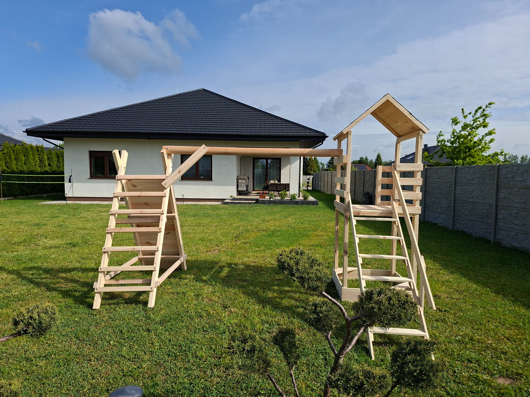 Nowy, drewniany plac zabaw z drabinkami na trawniku przed domem. Jasne drewno, błękitne niebo. Gotowy do zabawy, czekający na dzieci.
