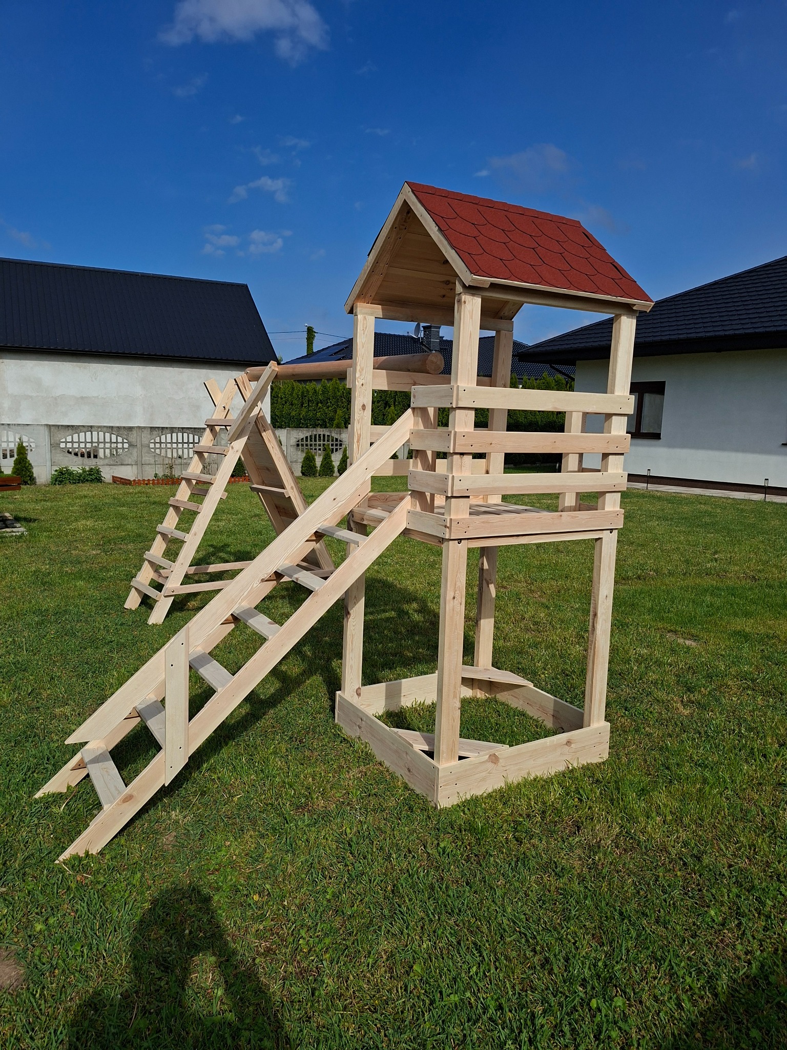 Drewniany plac zabaw z wieżą i schodami na trawniku, z domami w tle, w słoneczny dzień. Czerwony dach wieżyczki. Naturalne drewno.