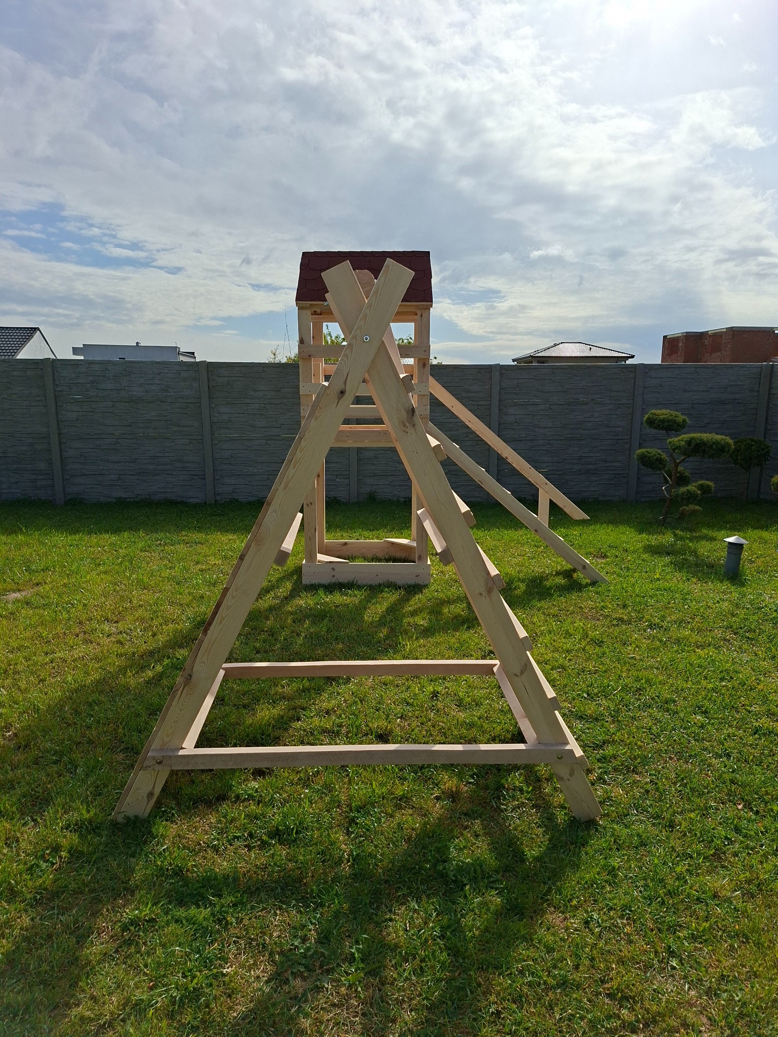 Drewniany plac zabaw z domkiem i zjeżdżalnią w ogrodzie, widok z dołu, jasne drewno, zielona trawa, błękitne niebo z chmurami w tle. Konstrukcja jeszcze nie pomalowana.