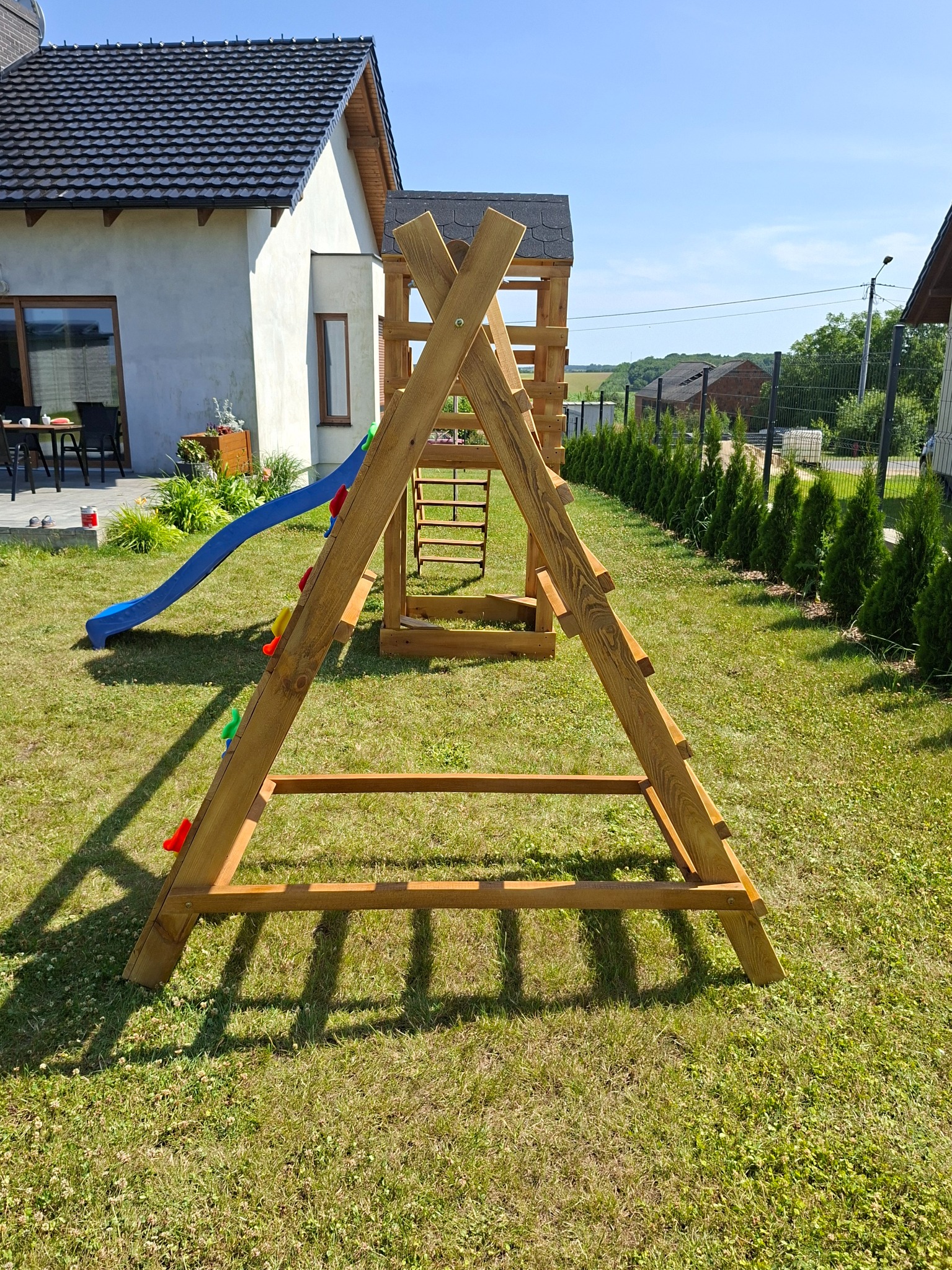 Drewniany plac zabaw dla dzieci z niebieską zjeżdżalnią, ustawiony na trawniku przed domem jednorodzinnym w słoneczny dzień. Widoczna drabinka i ścianka wspinaczkowa.