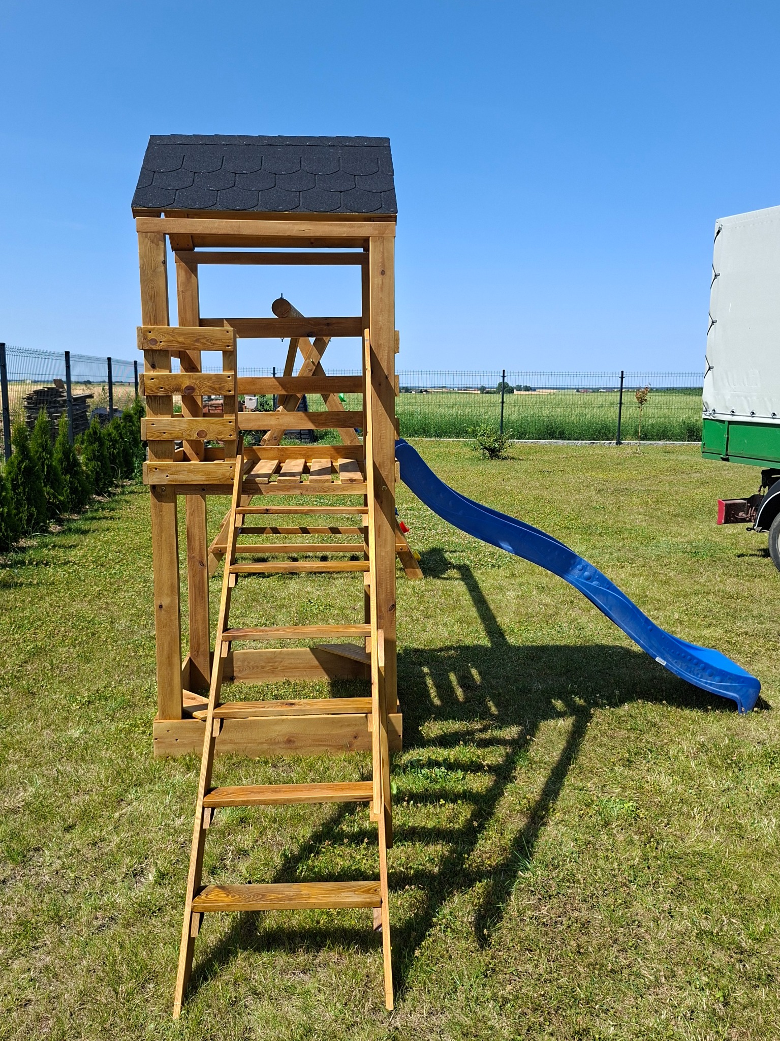 Drewniany plac zabaw z niebieską zjeżdżalnią, drabinką i małym domkiem na trawie, pod błękitnym niebem. Widoczny fragment przyczepy z plandeką w tle.