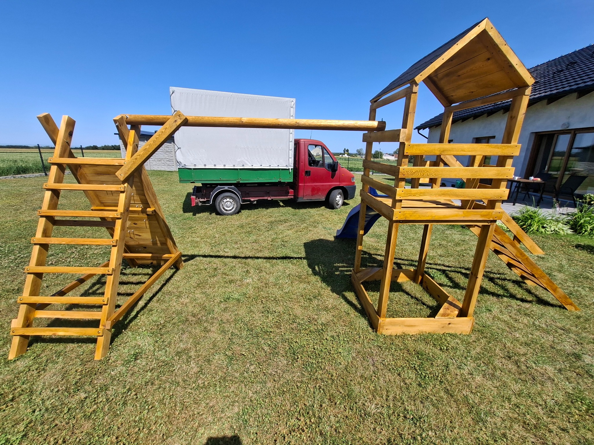 Drewniany plac zabaw z drabinkami i zjeżdżalnią na trawniku, w tle czerwony samochód dostawczy i budynek mieszkalny. Słoneczny dzień, błękitne niebo.