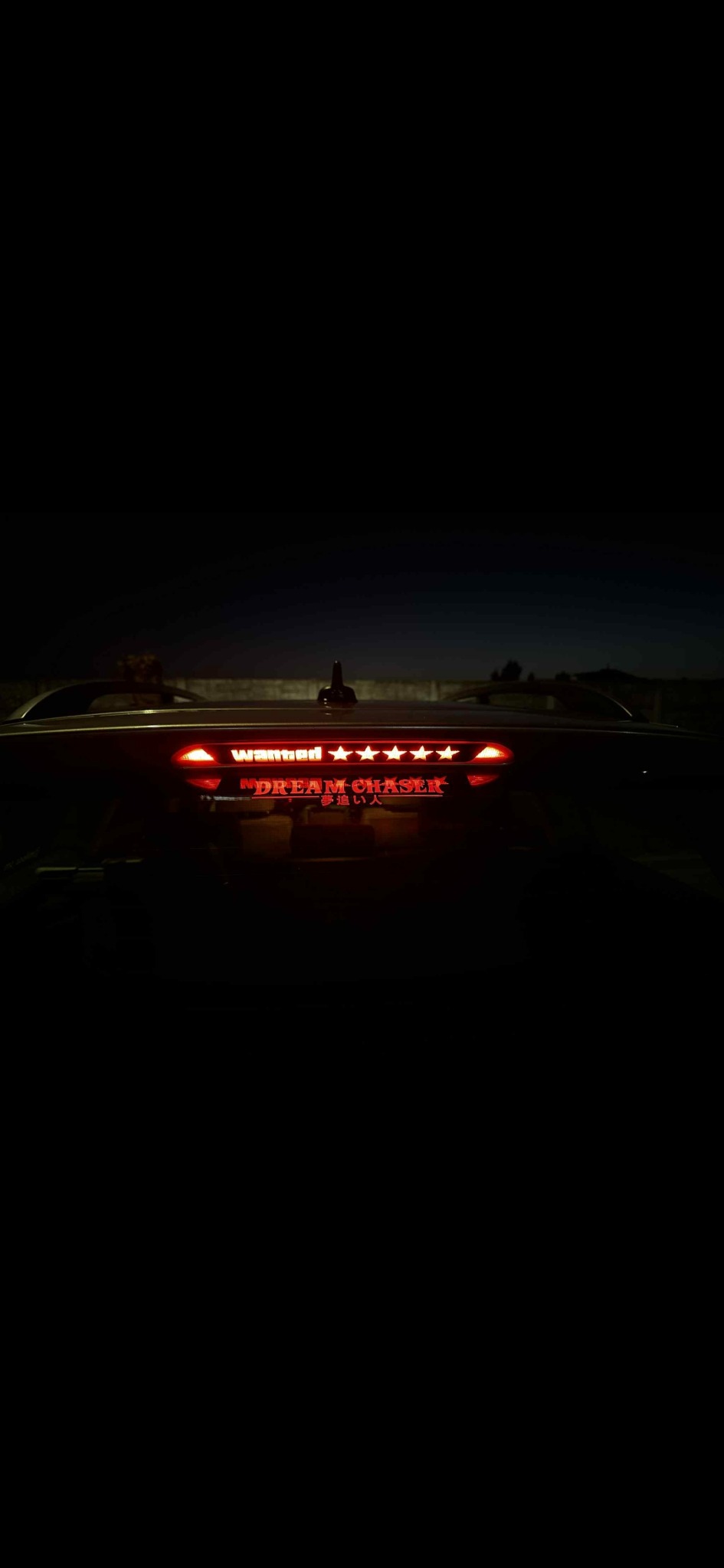 Tylna szyba auta z naklejkami 'Wanted' i 'Dream Chaser' podświetlonymi światłem stopu. Nocne ujęcie, widoczne światła i sylwetka pojazdu.