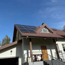 Dom z panelami słonecznymi na brązowym dachu, widok z dołu na tle błękitnego nieba. Detal: nietypowe okno trójkątne na poddaszu.