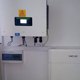 Biały inwerter Sofar z żółtą naklejką ostrzegawczą, zamontowany na ścianie nad rozdzielnicą elektryczną i akumulatorem. Widoczne kable i przewody.