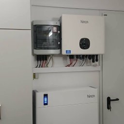 Zamontowany zestaw: rozdzielnica, inwerter i akumulator energii z niebieskim wskaźnikiem na białej ścianie. Widoczne przewody i elementy instalacji.