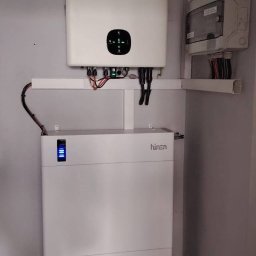 Biały inwerter i akumulator Hinen zamontowane na ścianie, z widocznymi kablami i skrzynką elektryczną. Instalacja w pomieszczeniu o jasnych ścianach.