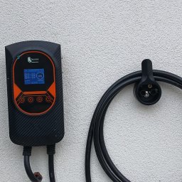 Czarna stacja ładowania Feyree z pomarańczowymi akcentami, zamontowana na białej ścianie obok uchwytu na kabel z wtyczką. Widoczny ekran z danymi ładowania.