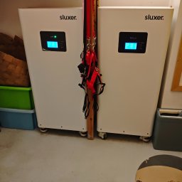 MDM Fotowoltaika - Dwa białe akumulatory energii z wyświetlaczami marki Sluxer, ustawione obok siebie w pomieszczeniu. Na ścianie zawieszony czerwony sprzęt wspinaczkowy.