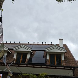 MDM Fotowoltaika - Instalacja fotowoltaiczna na dachu domu z brązową dachówką, widok z dołu, dekoracje z flag na pierwszym planie, pochmurne niebo w tle.
