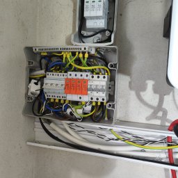 MDM Fotowoltaika - Otwarta skrzynka elektryczna z bezpiecznikami i przewodami na szarej ścianie, instalacja elektryczna zabezpieczeń przepięciowych w systemie OZE.