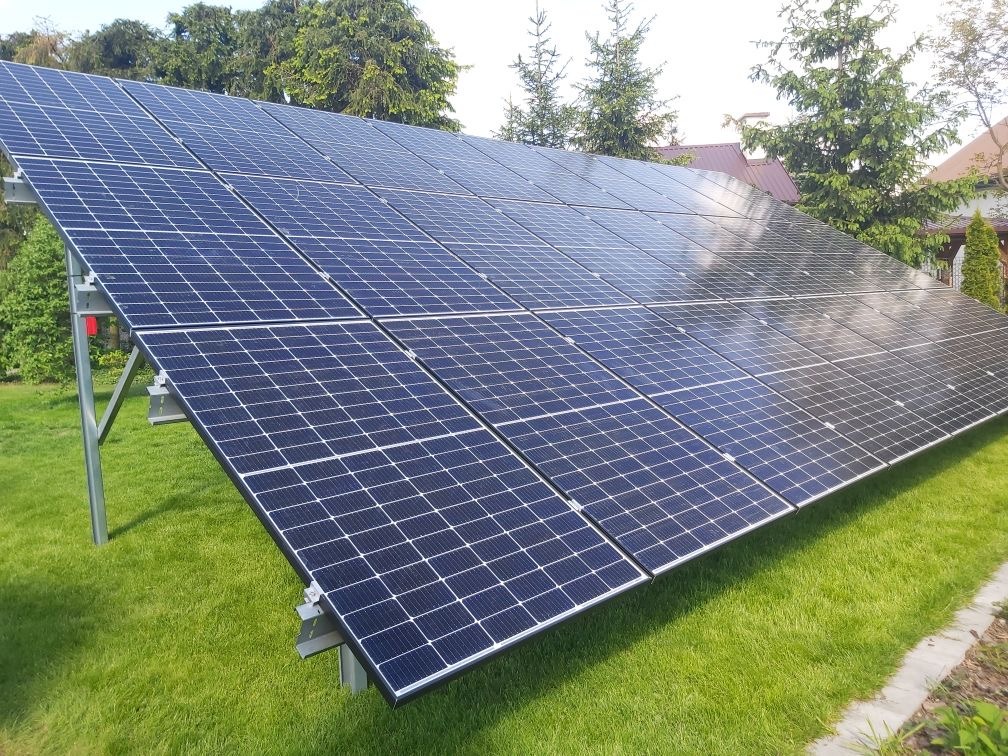 Instalacja fotowoltaiczna na trawniku, panele ustawione pod kątem, widoczne wsporniki montażowe. Tło: drzewa, fragment budynku. Słoneczny dzień.