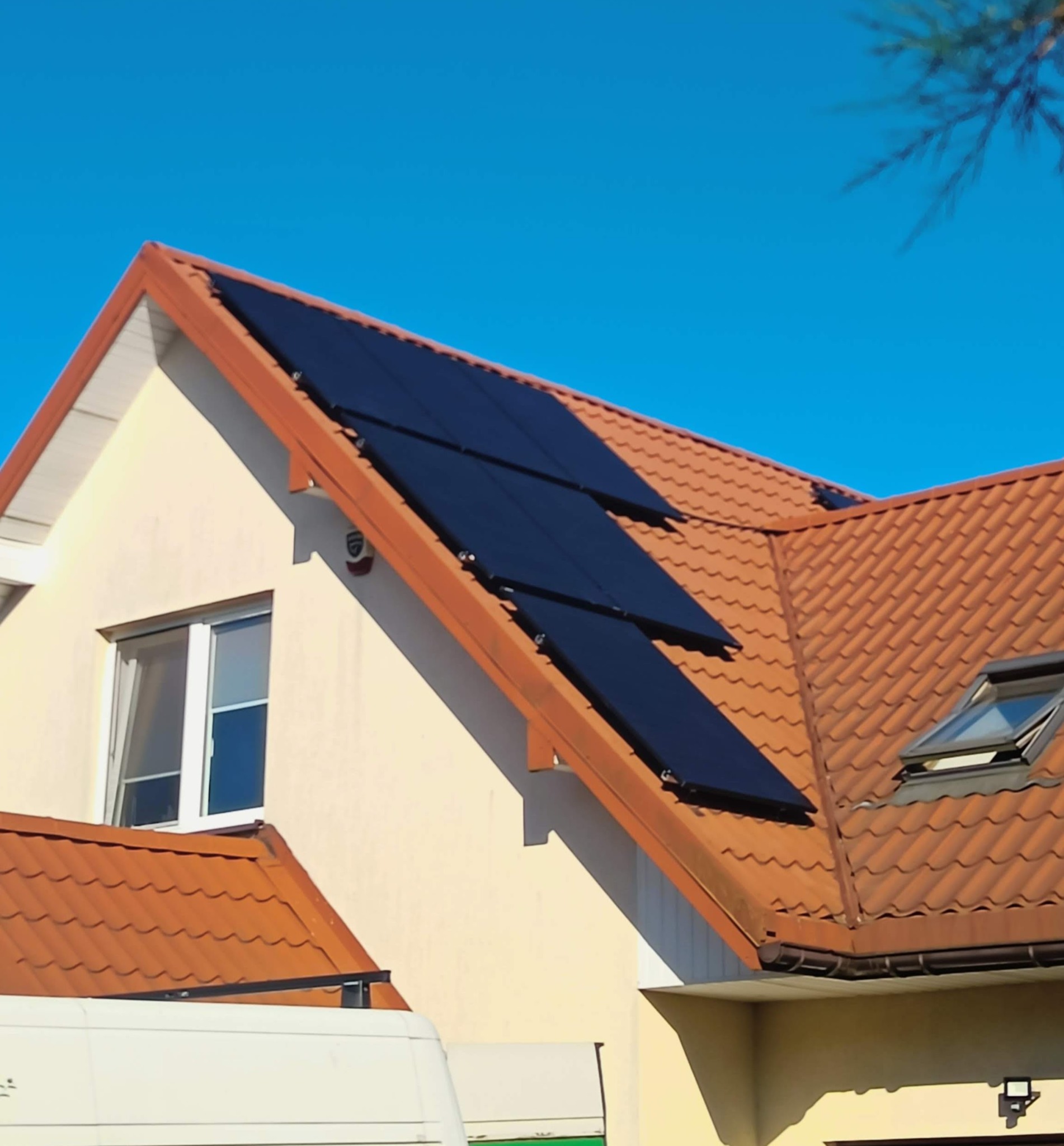 Panele słoneczne na dachu domu z dachówką ceramiczną w słoneczny dzień. Widoczny fragment białego busa i okno dachowe.