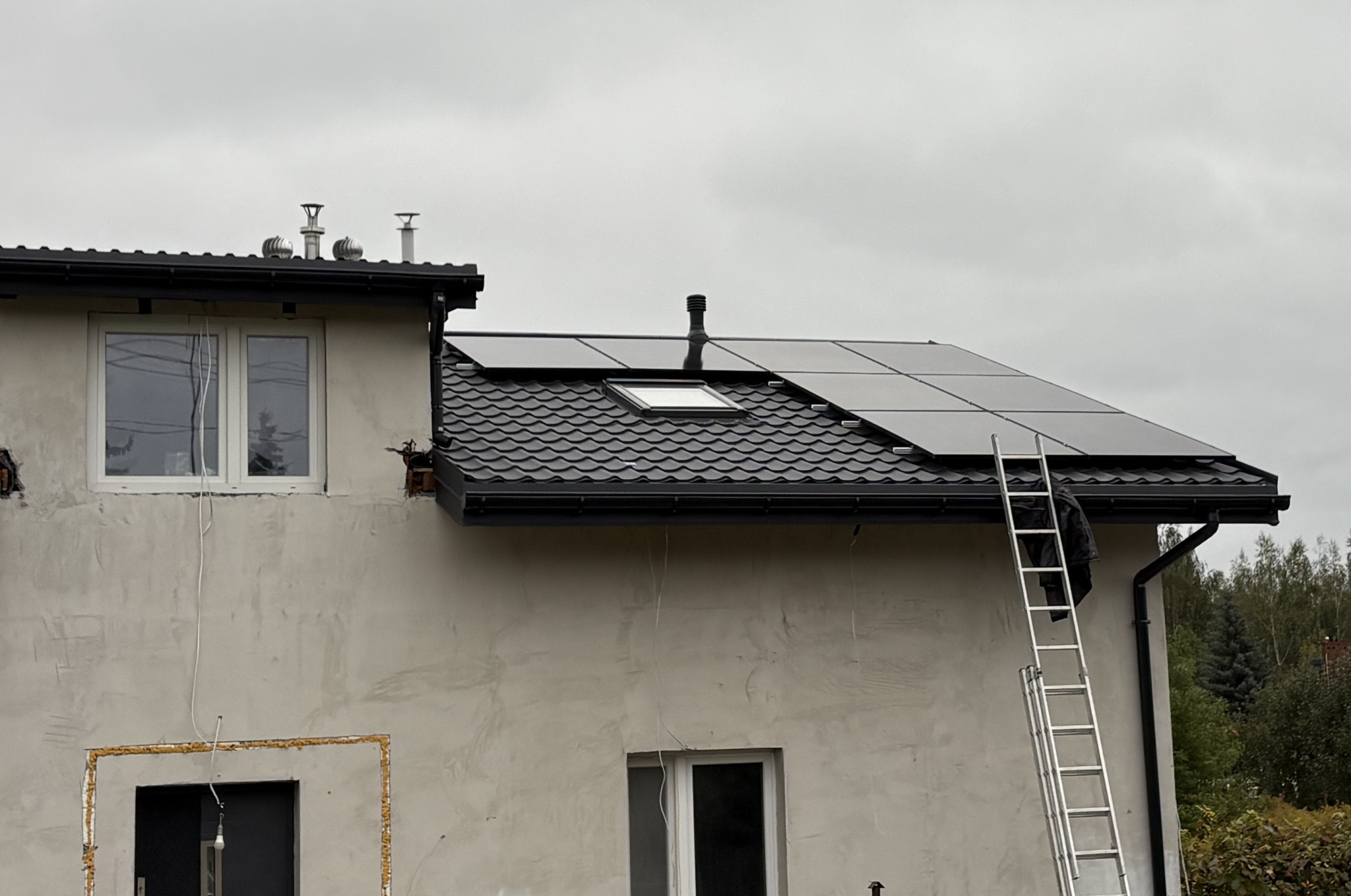 Panele słoneczne na dachu domu z czarną dachówką, drabina oparta o rynnę, widoczne okno dachowe i fragment elewacji w trakcie wykończenia.