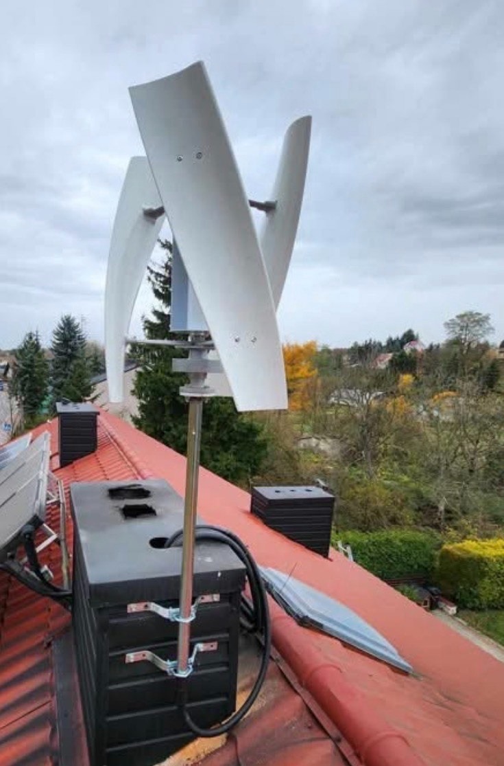 Turbina wiatrowa pionowa zamontowana na dachu budynku z czerwoną dachówką, widok z góry. W tle drzewa i zabudowa mieszkalna.