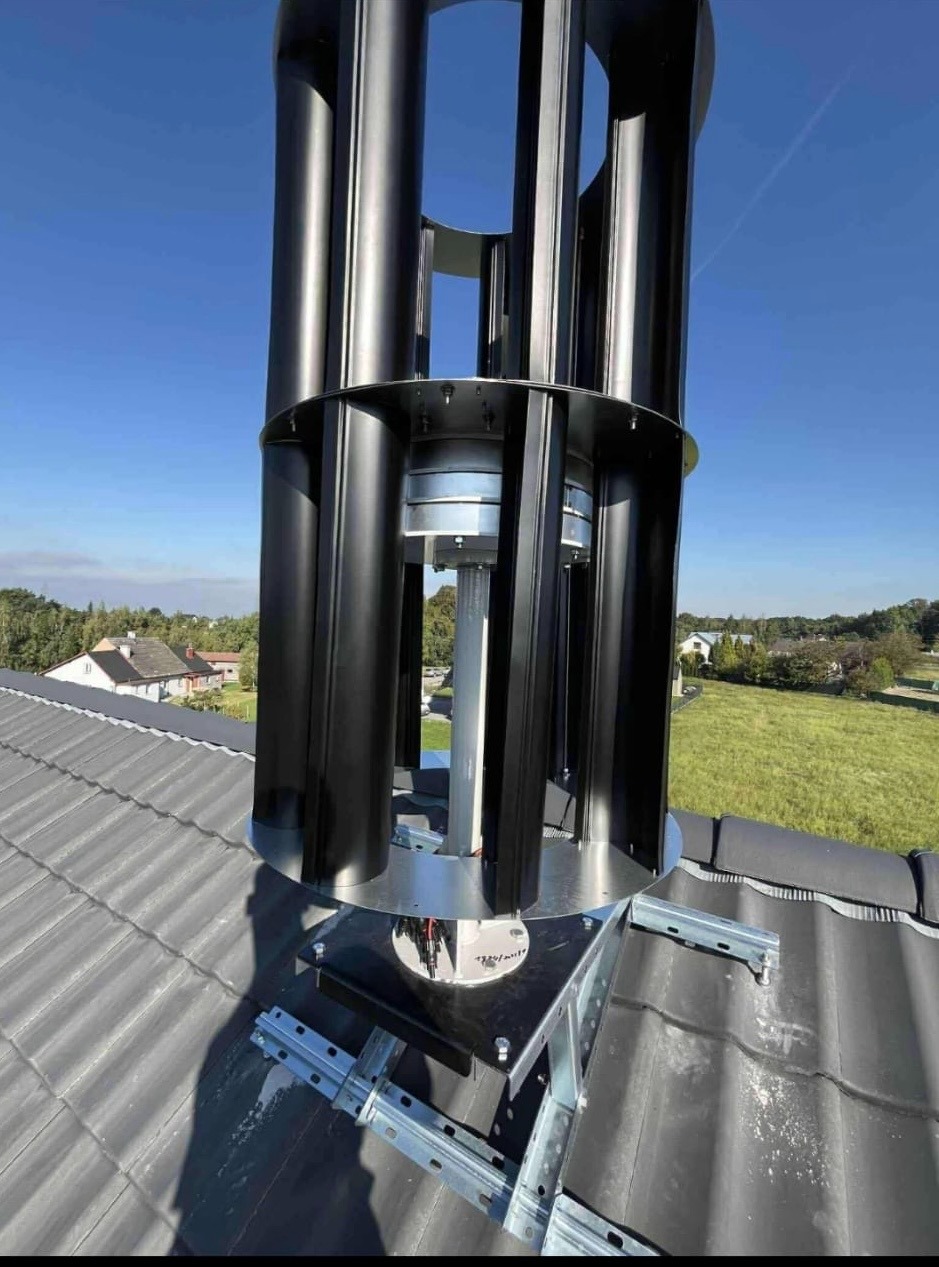 Turbina wiatrowa pionowa, zamontowana na szarym dachu domu jednorodzinnego, na tle błękitnego nieba i zieleni. Widoczne elementy montażowe i konstrukcja turbiny.