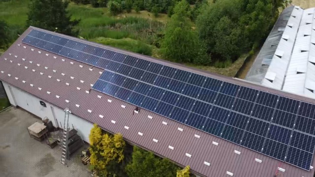 Panele słoneczne na brązowym dachu budynku gospodarczego, widok z góry. Obok szklarnia, w tle zieleń drzew. Drabina oparta o ścianę budynku.
