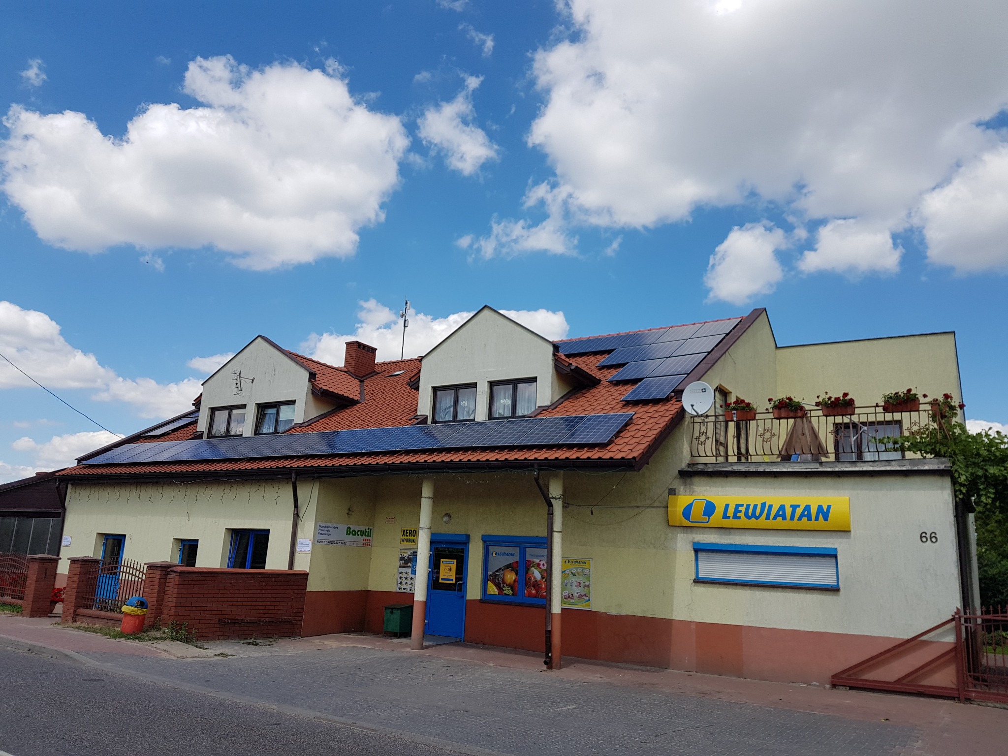 Budynek sklepu Lewiatan z panelami słonecznymi na dachu, widok z ulicy w słoneczny dzień. Czerwona dachówka i kwiaty na balkonie dodają uroku.