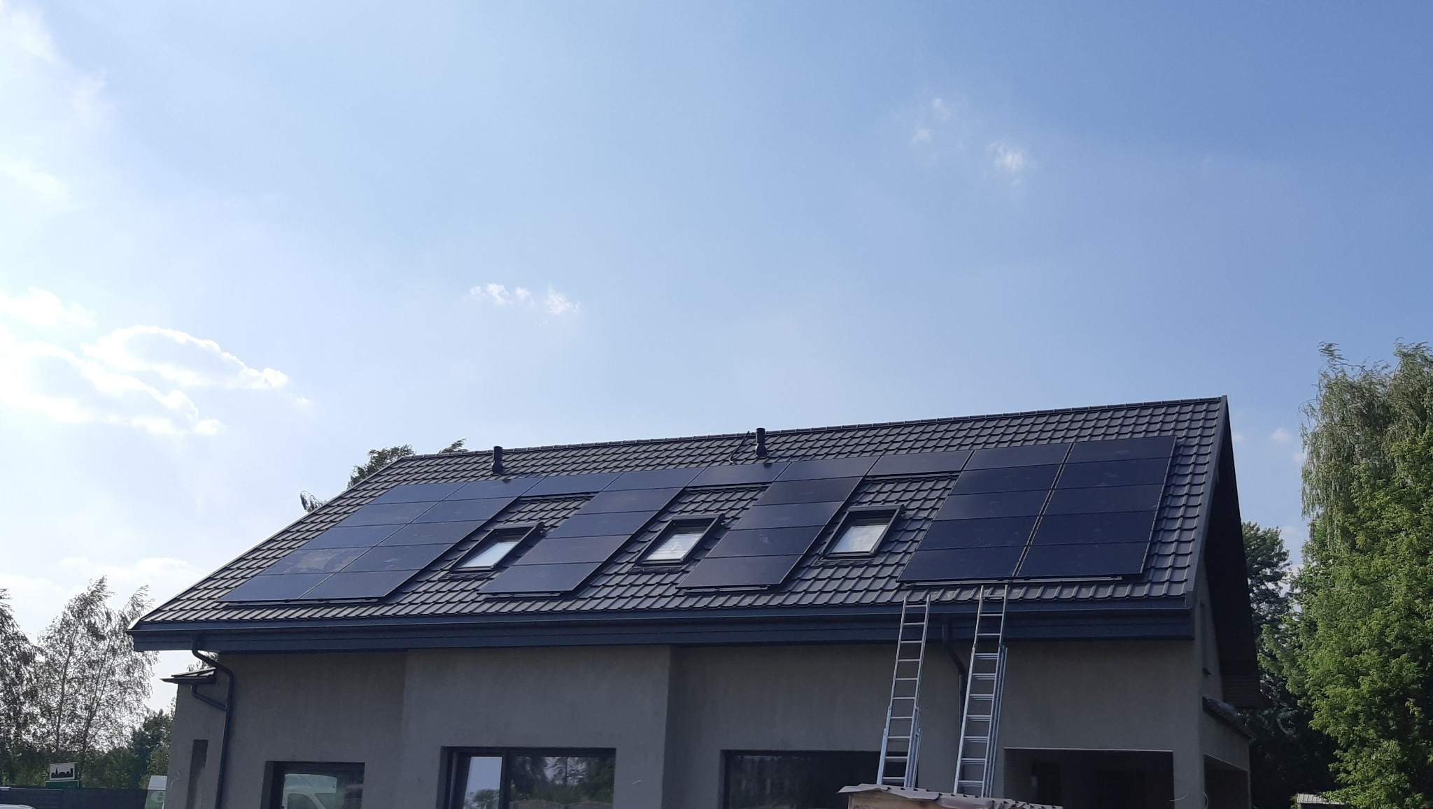 Panele słoneczne na dachu domu jednorodzinnego z oknami dachowymi, drabina oparta o dach. Błękitne niebo w tle. Widok z dołu, lekko z prawej strony.