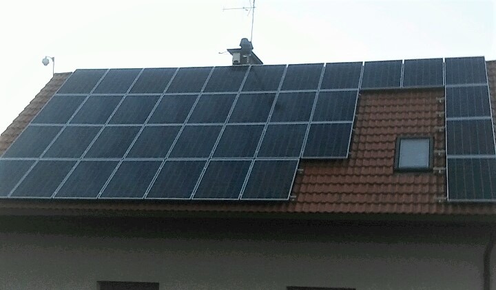 Panele słoneczne na dachu domu z czerwoną dachówką, widoczne okno dachowe i antena. Montaż paneli częściowo ukończony, widoczne mocowania.