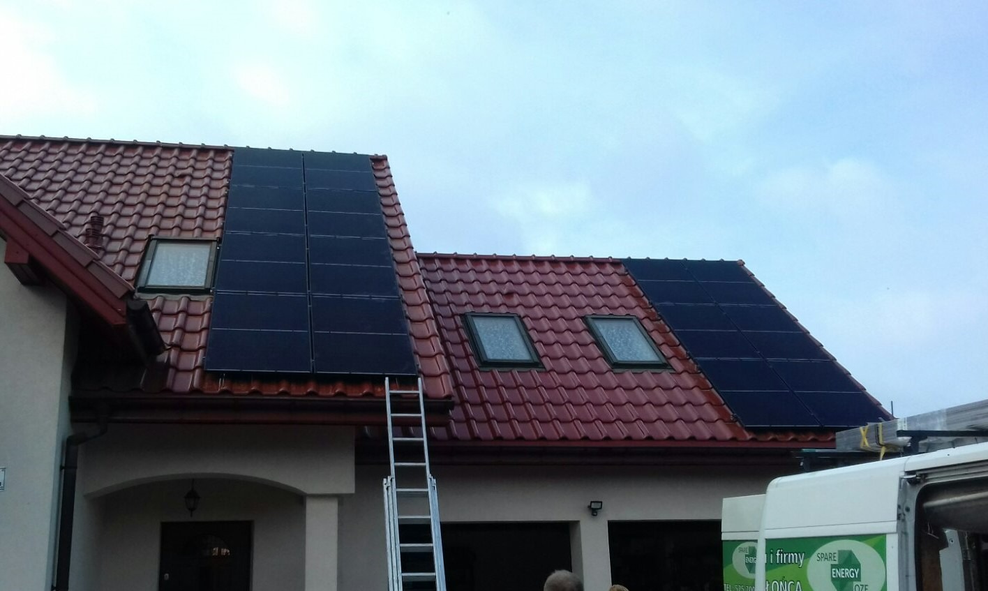 Częściowy widok dachu z czerwoną dachówką, na którym zamontowano panele fotowoltaiczne. Widoczna drabina oparta o dach i fragment białego samochodu dostawczego.