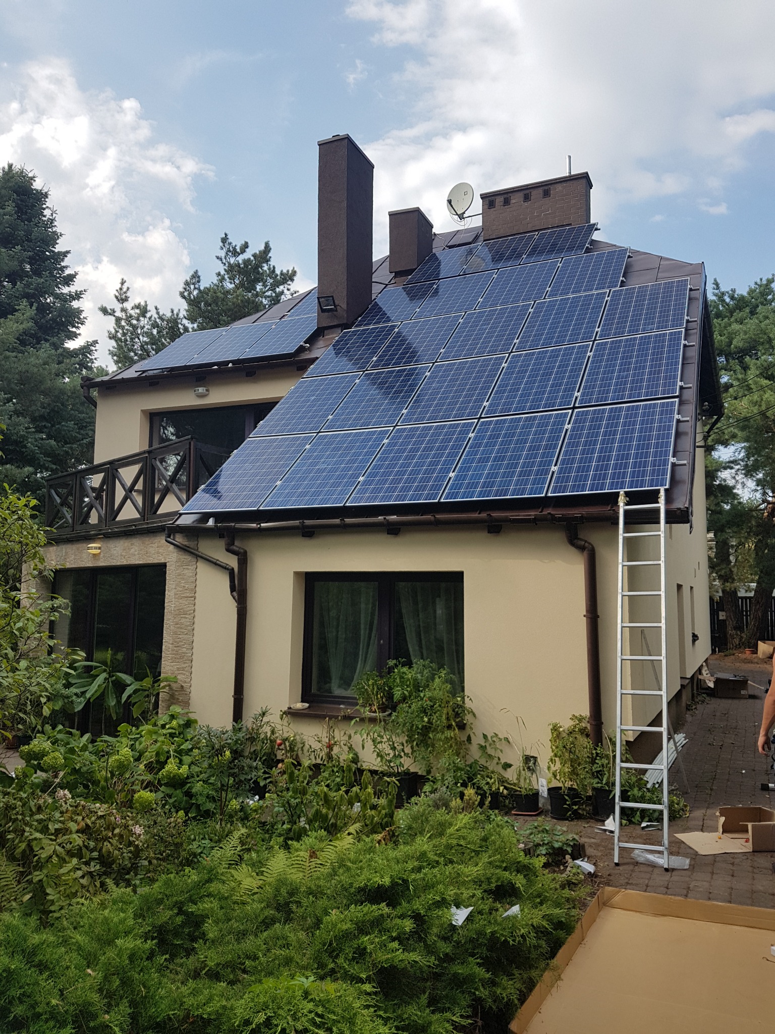 Dom jednorodzinny z panelami słonecznymi na dachu, drabina oparta o ścianę, bujna roślinność w ogrodzie. Widok z dołu, częściowo pochmurne niebo.