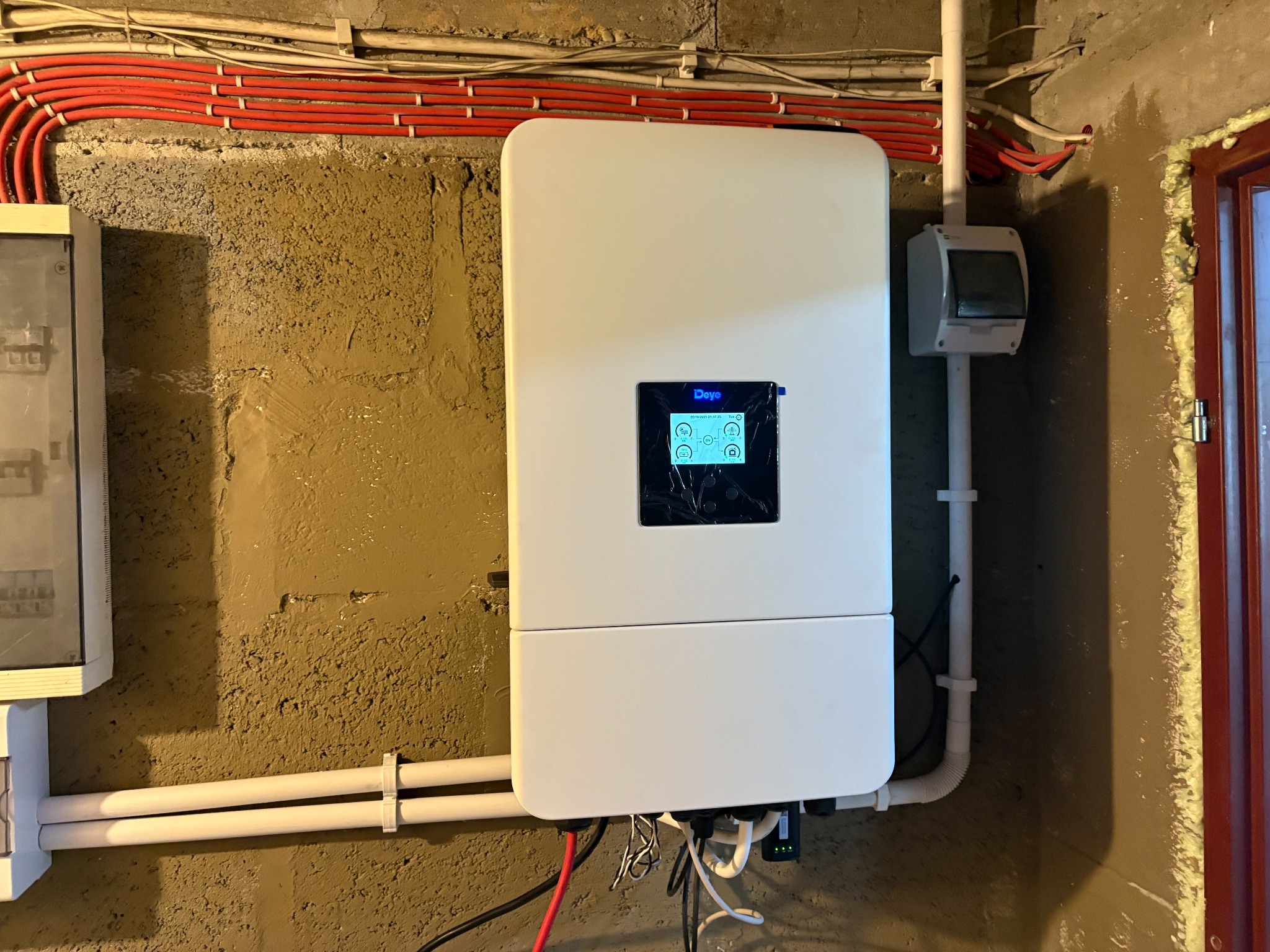 Zamontowany inwerter na ścianie z widocznymi przewodami i skrzynką elektryczną. Instalacja wewnątrz budynku z surowym wykończeniem ściany.