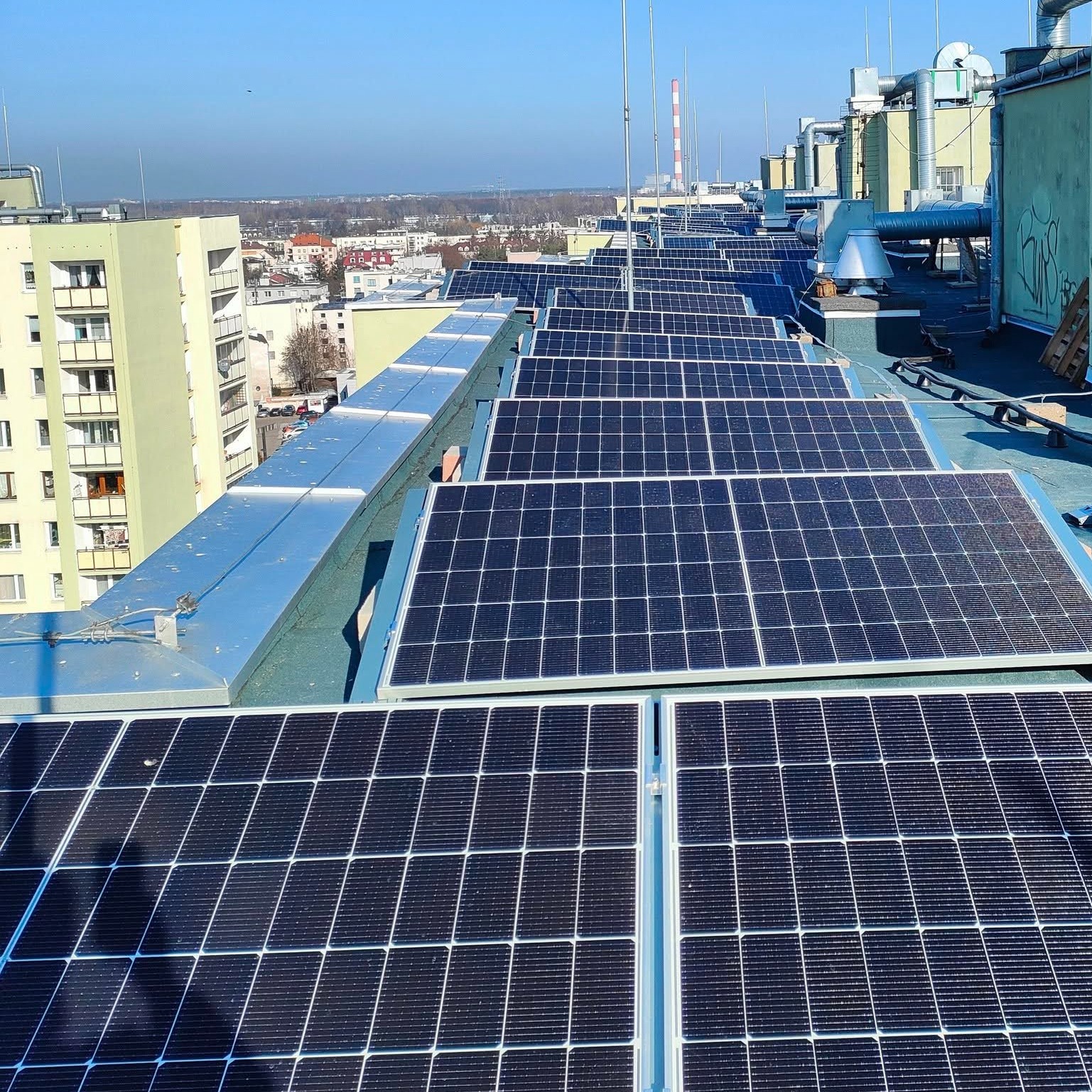 Rząd paneli słonecznych na dachu budynku, z widokiem na miasto w tle. Panele ułożone równo, widać konstrukcję montażową. Słoneczny dzień.