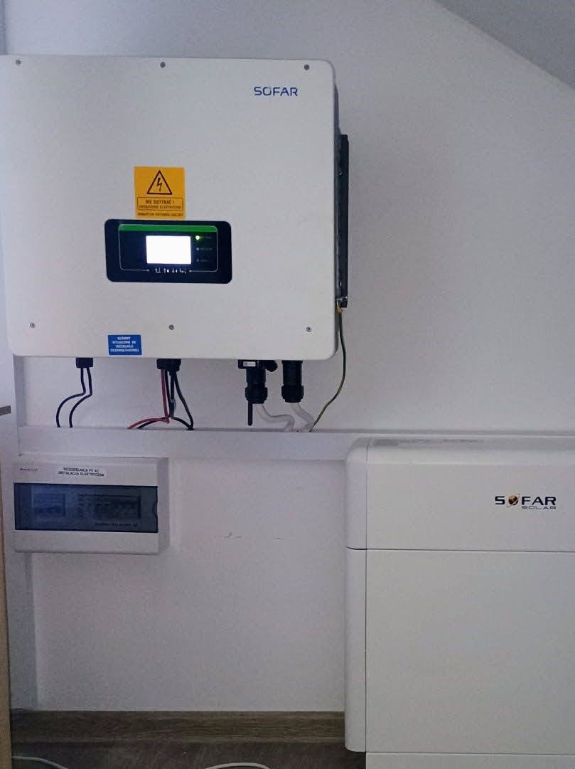 Biały inwerter Sofar z żółtą naklejką ostrzegawczą, zamontowany na ścianie nad rozdzielnicą elektryczną i akumulatorem. Widoczne kable i przewody.