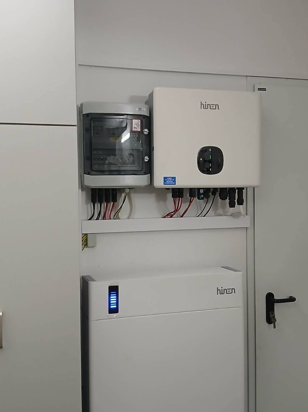 Zamontowany zestaw: rozdzielnica, inwerter i akumulator energii z niebieskim wskaźnikiem na białej ścianie. Widoczne przewody i elementy instalacji.