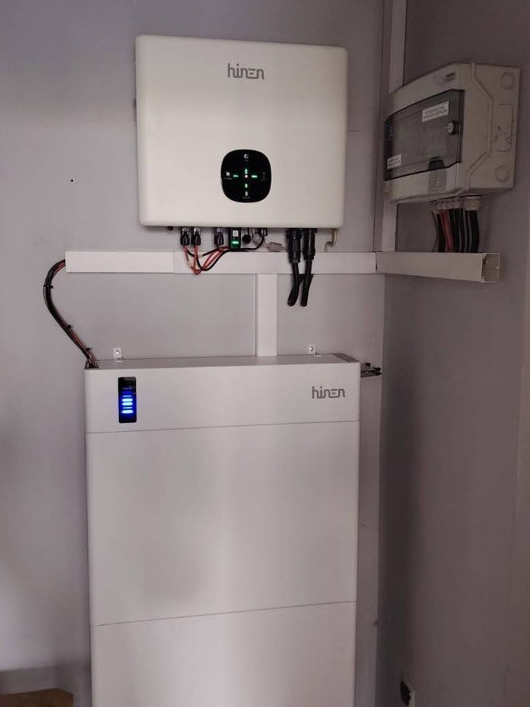 Biały inwerter i akumulator Hinen zamontowane na ścianie, z widocznymi kablami i skrzynką elektryczną. Instalacja w pomieszczeniu o jasnych ścianach.