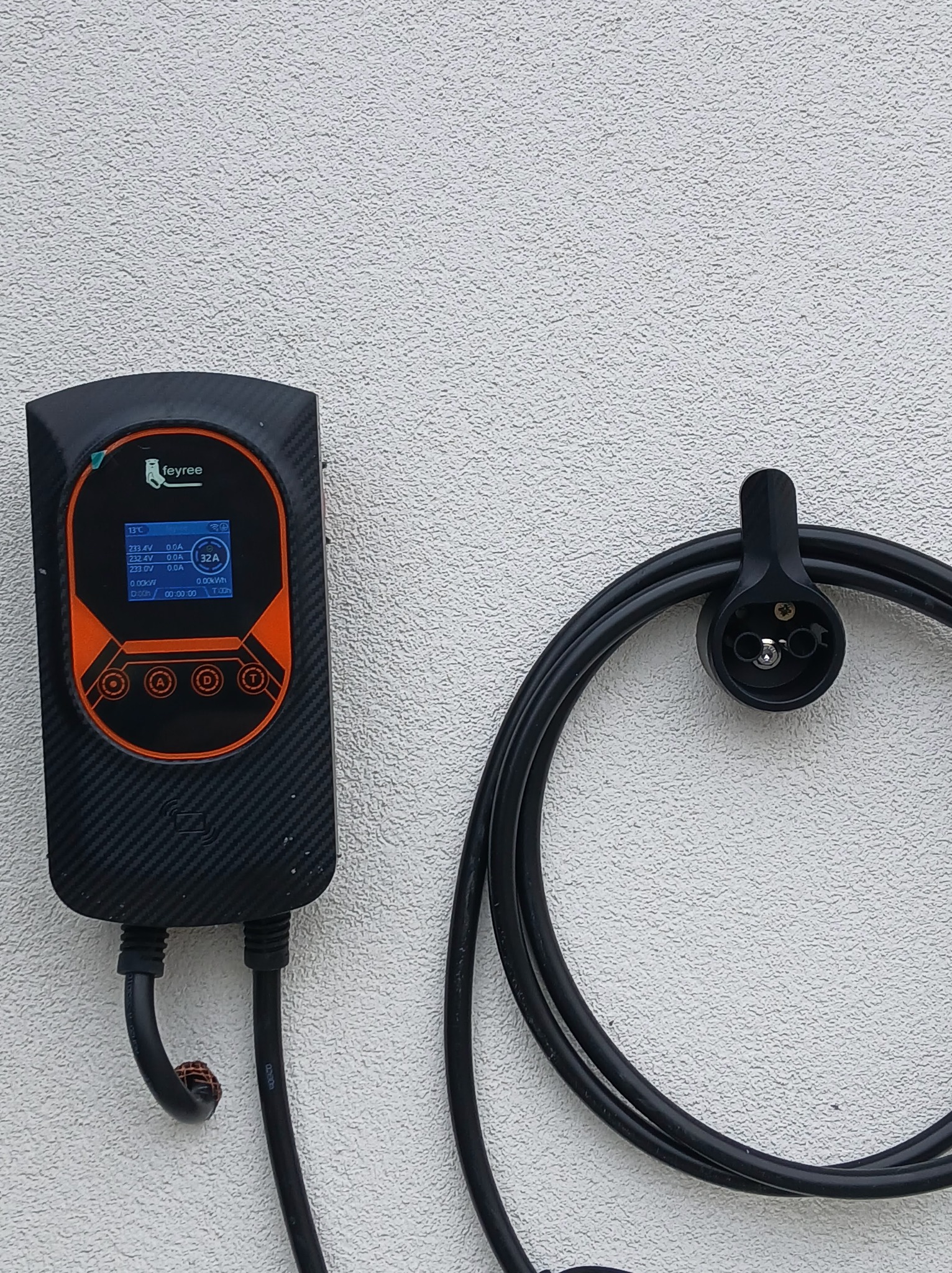 Czarna stacja ładowania Feyree z pomarańczowymi akcentami, zamontowana na białej ścianie obok uchwytu na kabel z wtyczką. Widoczny ekran z danymi ładowania.