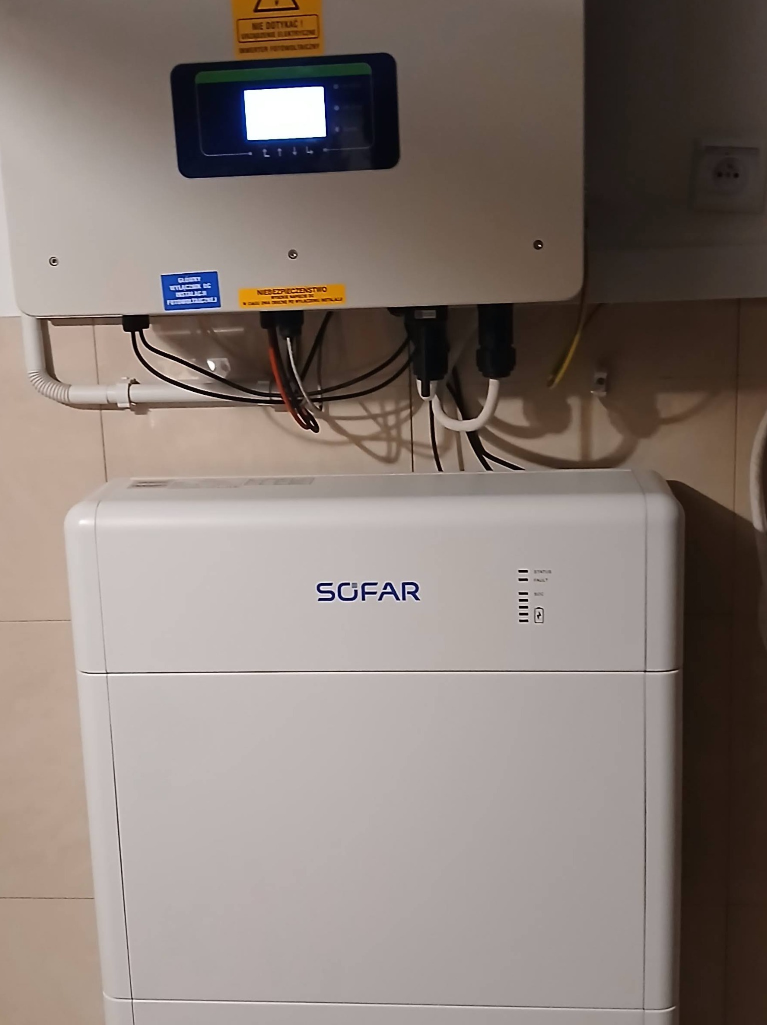 Biały inwerter Sofar z podłączonymi kablami, zamontowany na ścianie. Widoczne naklejki ostrzegawcze i gniazdko elektryczne. Instalacja wewnątrz budynku.