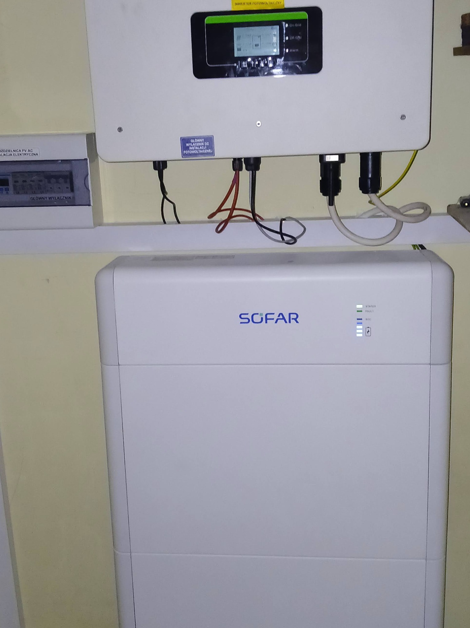 Biały inwerter Sofar z podłączonymi kablami, zamontowany na ścianie obok skrzynki elektrycznej. Widoczne diody statusu i wyświetlacz inwertera.