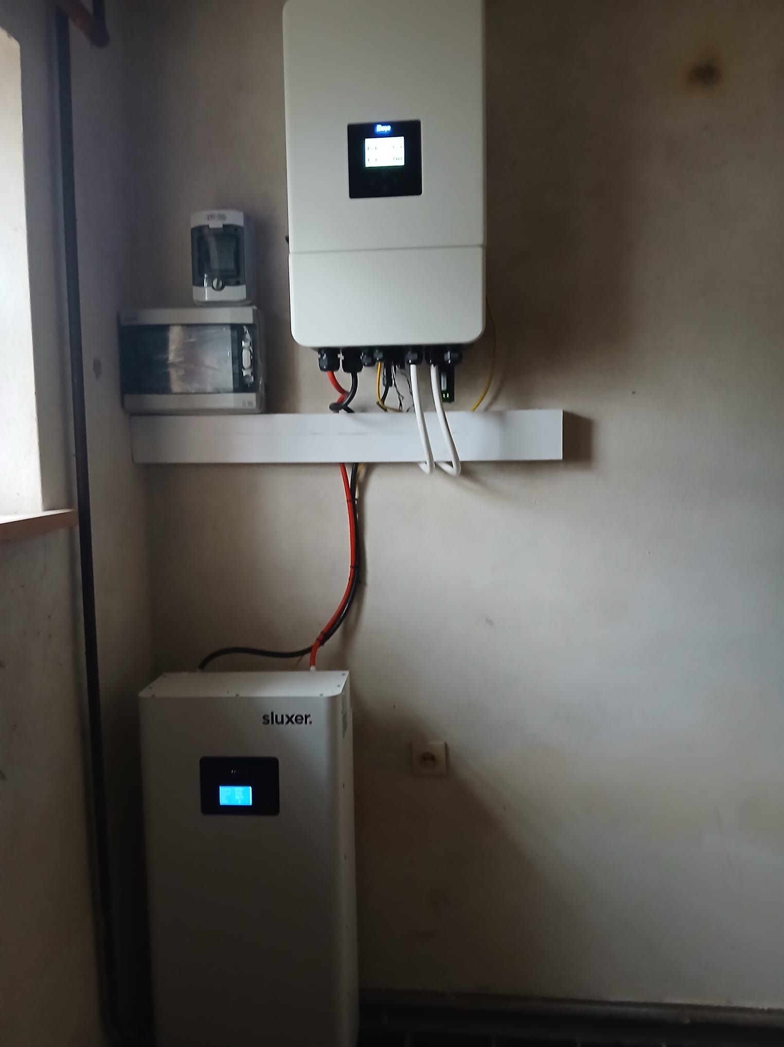 Zamontowany inwerter i akumulator marki Sluxer na ścianie wewnątrz budynku. Widoczne okablowanie i elementy instalacji.