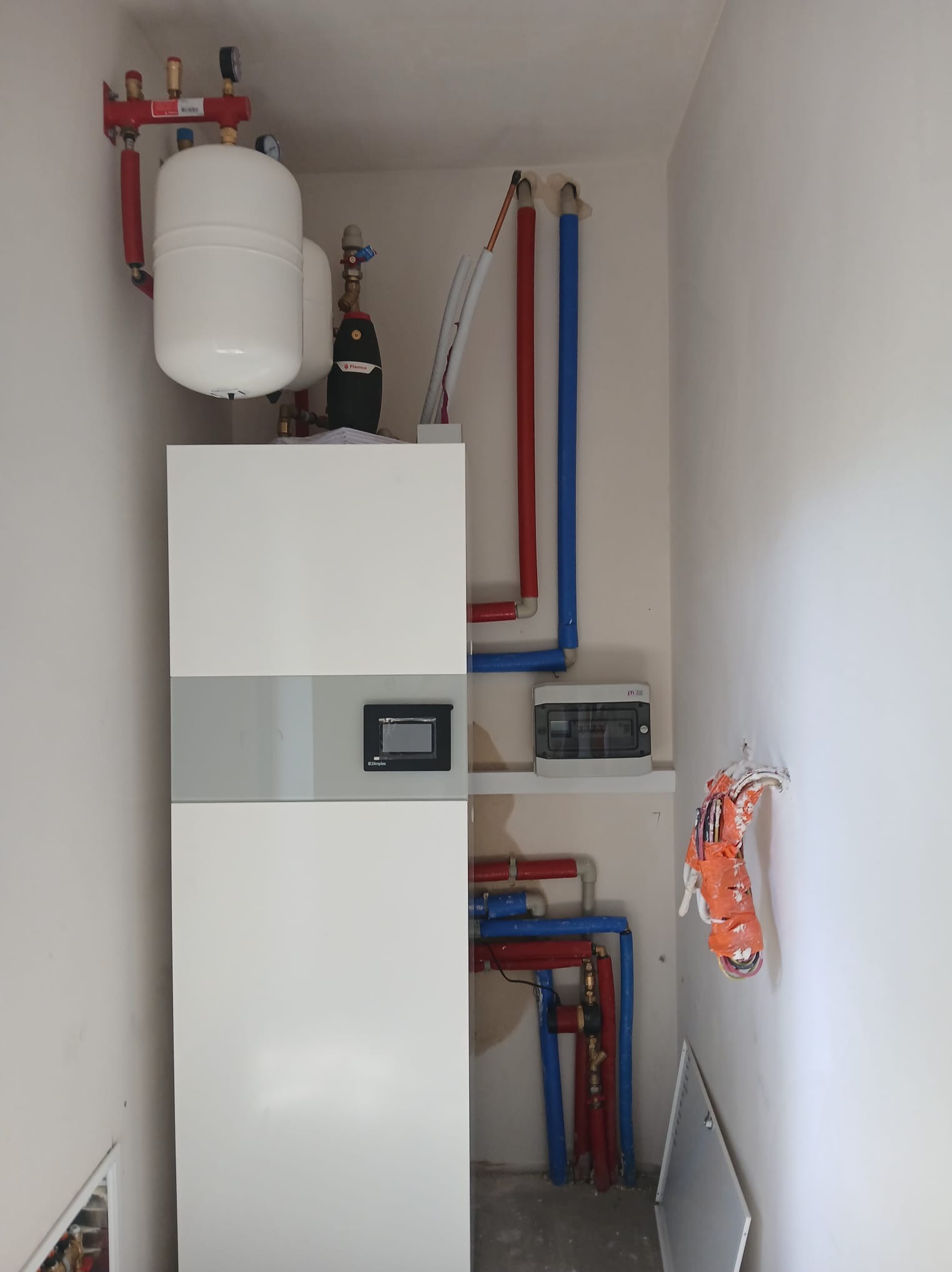 Wnętrze kotłowni z nowoczesnym, białym piecem, zbiornikiem wyrównawczym i instalacją hydrauliczną z rurami w kolorach czerwonym i niebieskim. Widoczna skrzynka elektryczna i wystające przewody.