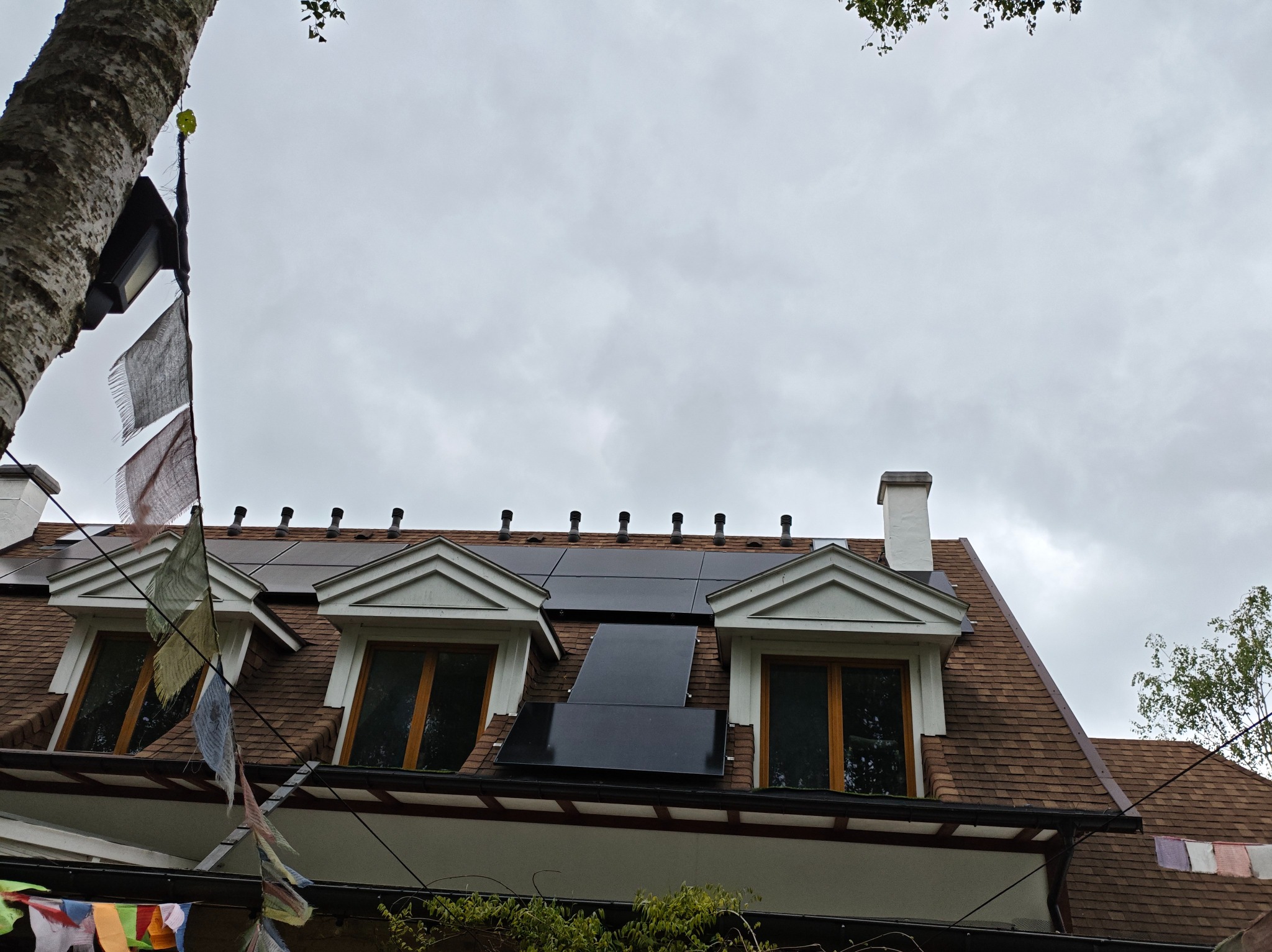 Instalacja fotowoltaiczna na dachu domu z brązową dachówką, widok z dołu, dekoracje z flag na pierwszym planie, pochmurne niebo w tle.