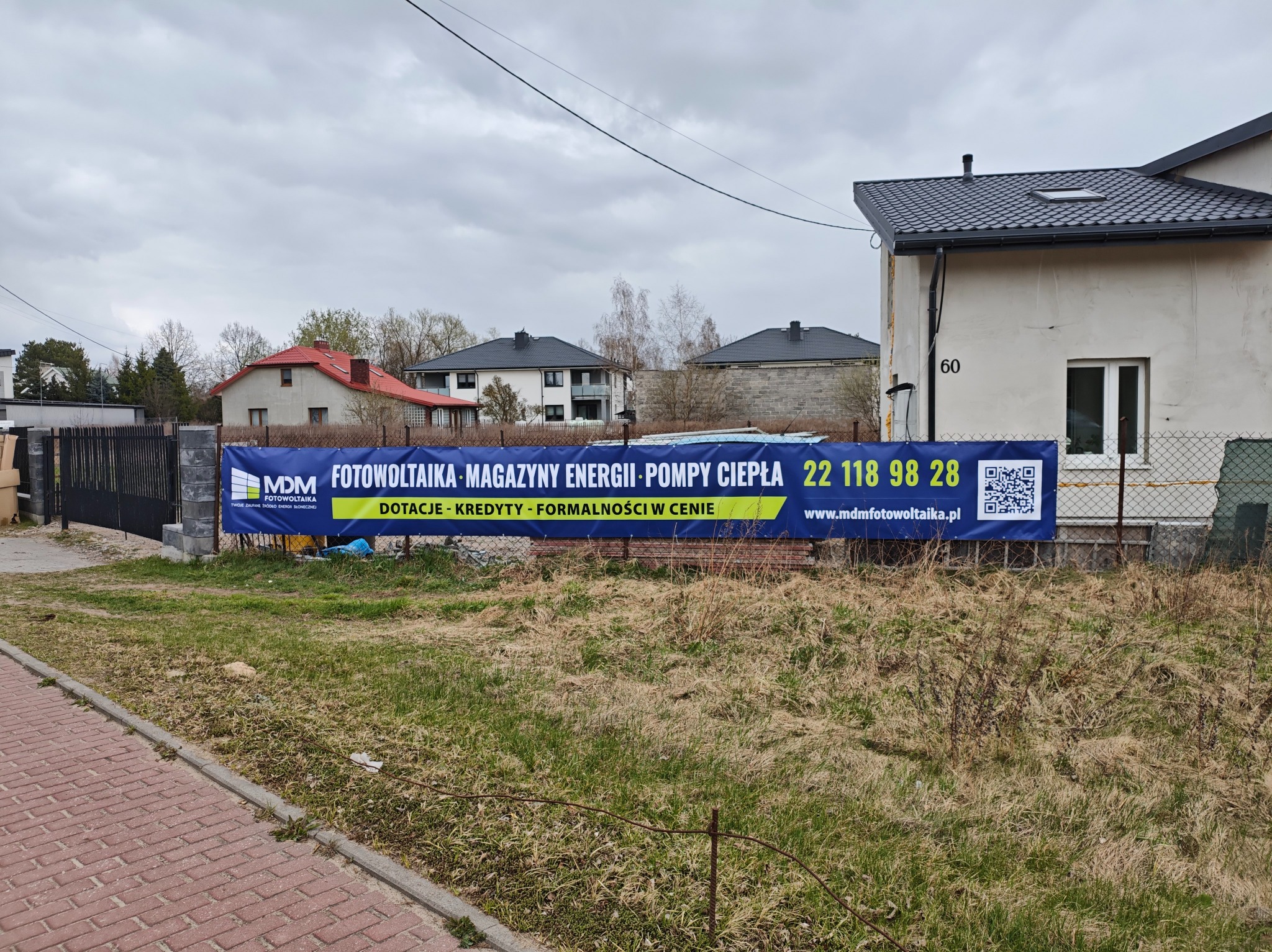 Niebieski baner reklamowy z ofertą na fotowoltaikę, magazyny energii i pompy ciepła, umieszczony na ogrodzeniu posesji z numerem 60 w tle.