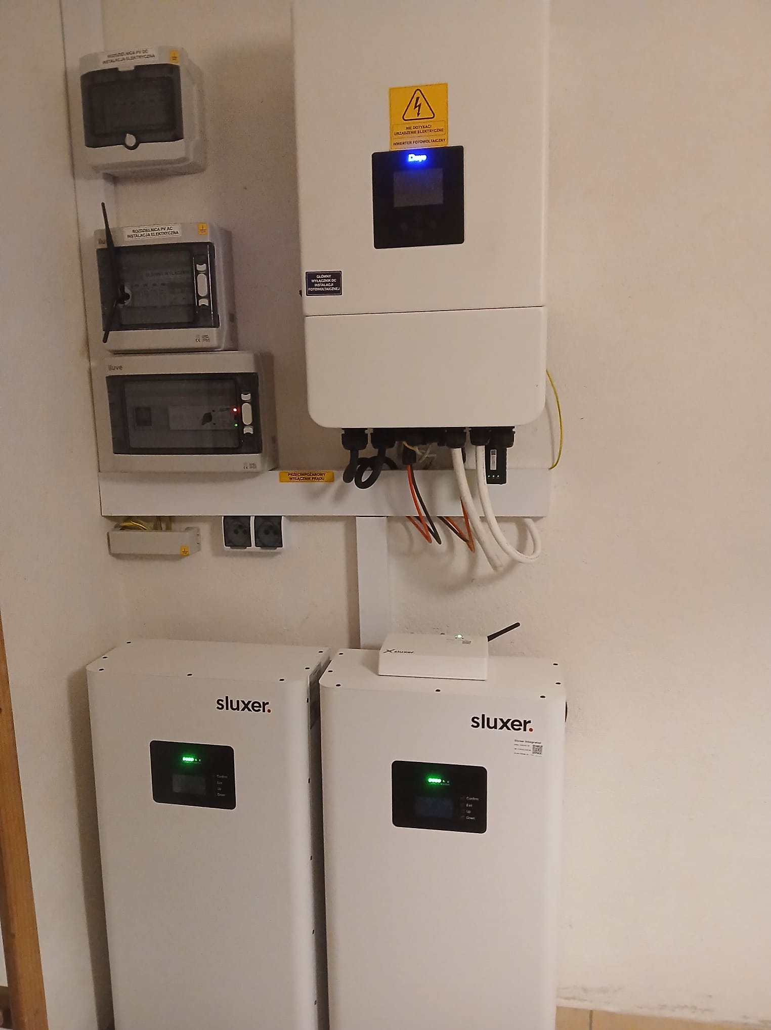 Instalacja z dwoma inwerterami Sluxer, rozdzielnicami i zabezpieczeniami. Widoczne okablowanie i elementy montażowe. Całość zamontowana na białej ścianie.