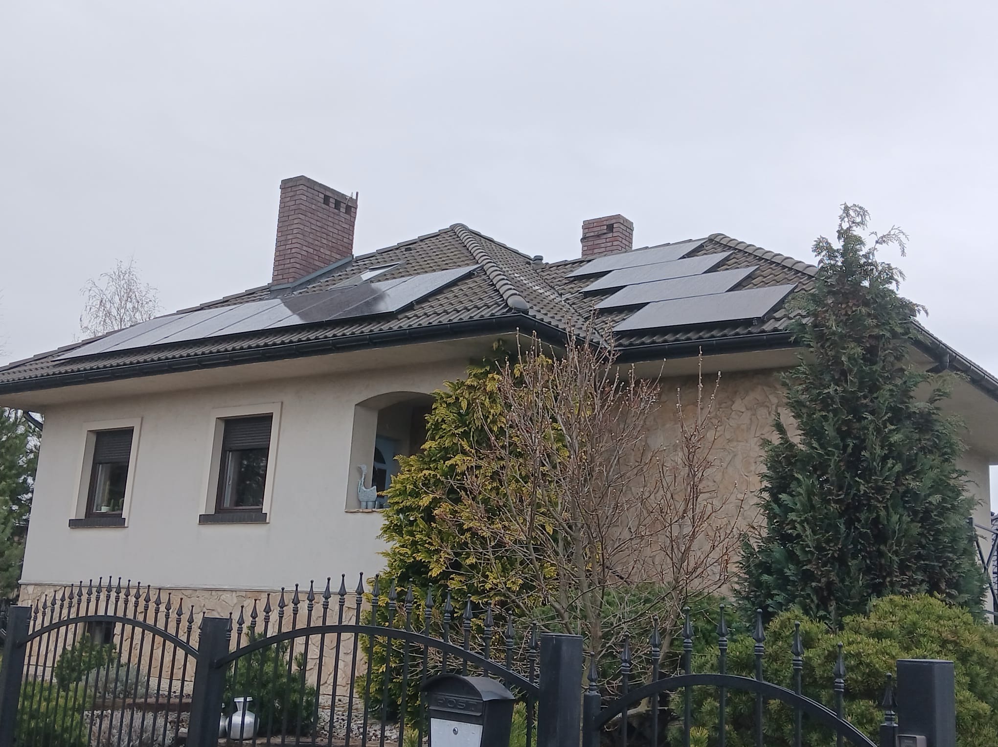 Dom jednorodzinny z panelami fotowoltaicznymi na dachu, widoczne kominy i ogrodzenie. Ujęcie z poziomu ulicy, pochmurne niebo.