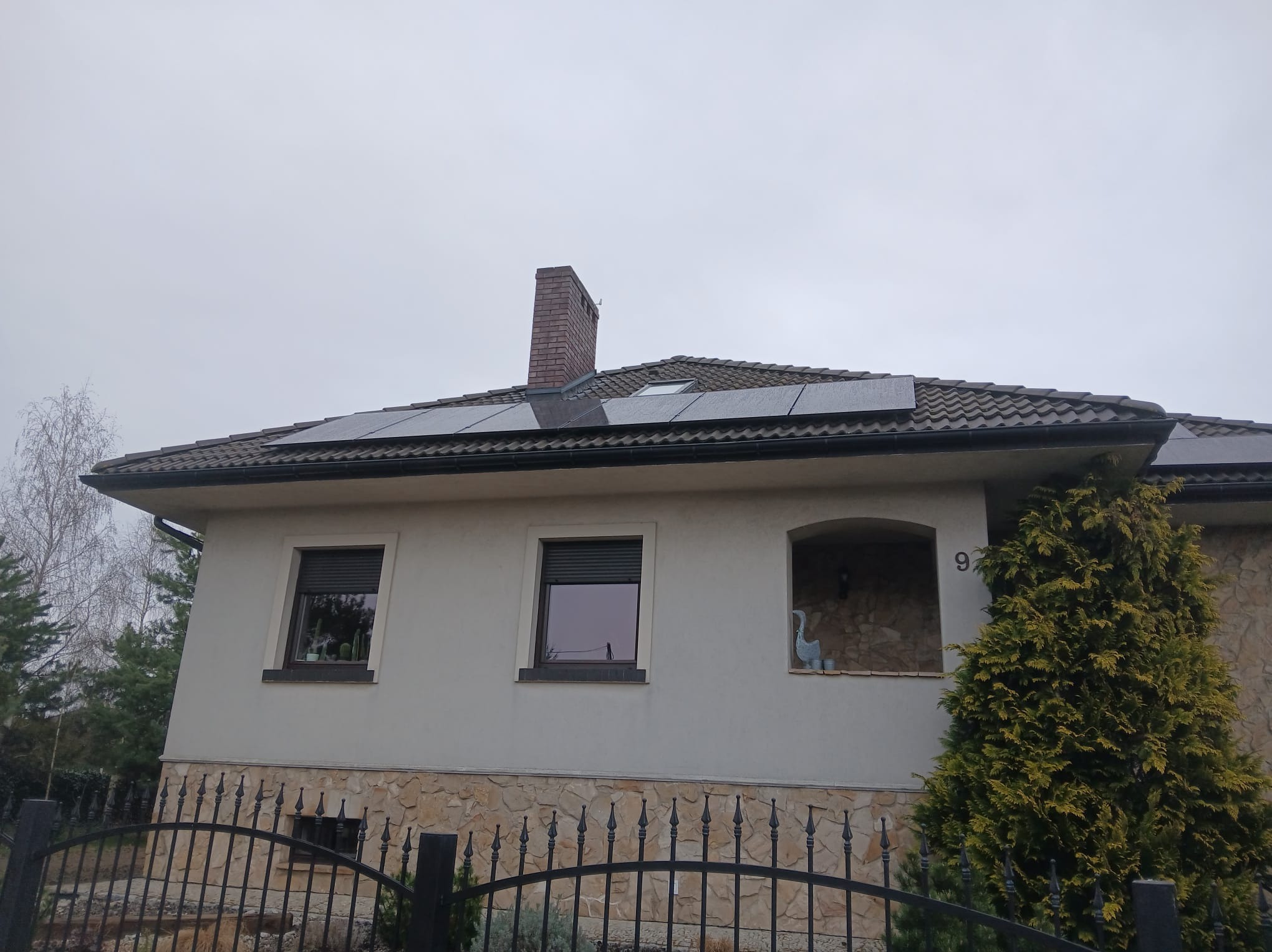 Dom jednorodzinny z panelami słonecznymi na dachu pokrytym dachówką, widok zza czarnego, metalowego ogrodzenia. Pochmurne niebo w tle.