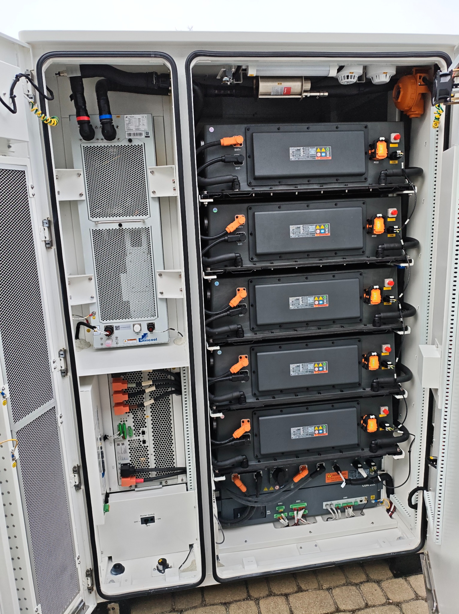 Otwarta szafa sterownicza z akumulatorami i elektroniką, widoczne kable, złącza i elementy chłodzenia. Biała obudowa, szara elektronika, pomarańczowe złącza.
