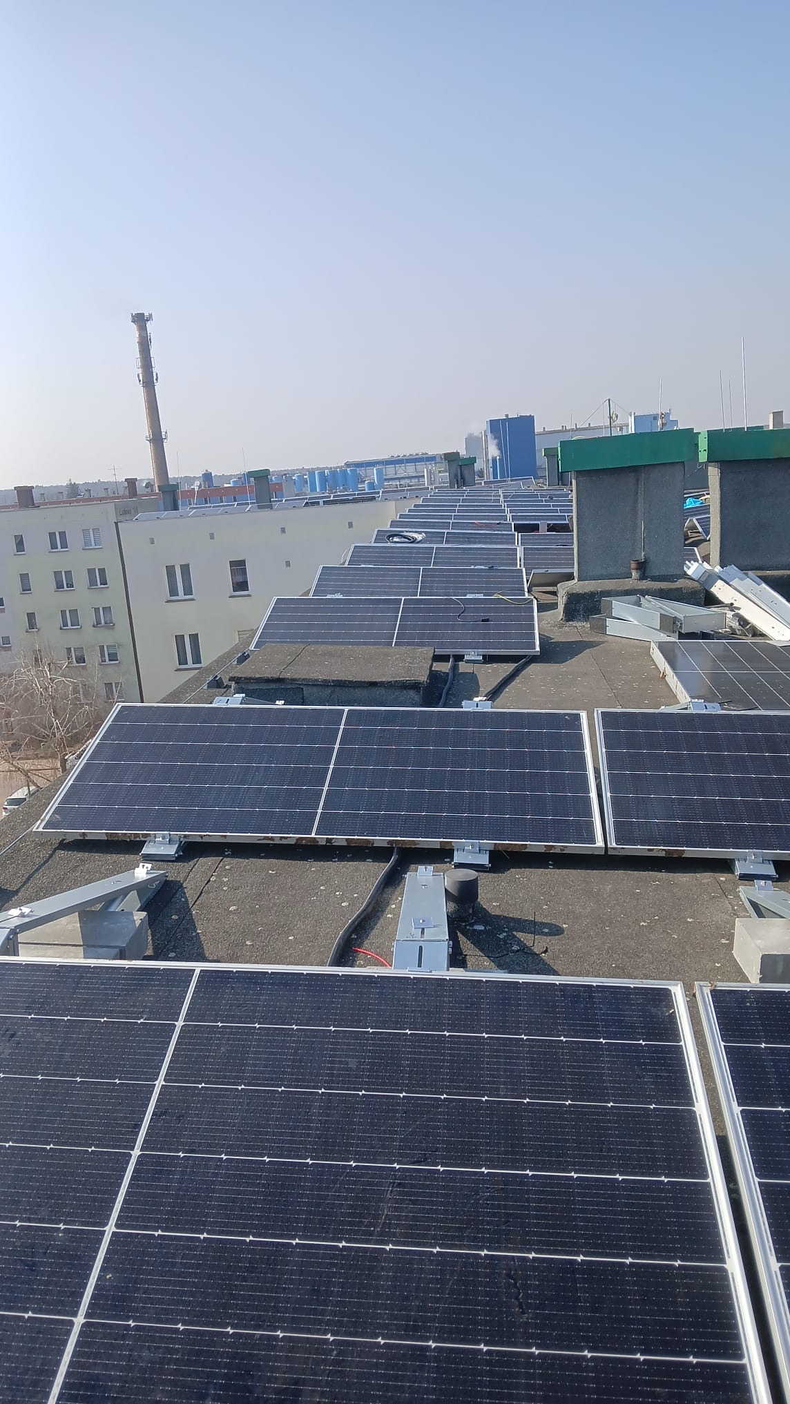 Rząd paneli słonecznych na płaskim dachu budynku, z kominem w tle i błękitnym niebem. Widoczne elementy montażowe i kable. Instalacja w trakcie lub tuż po zakończeniu.