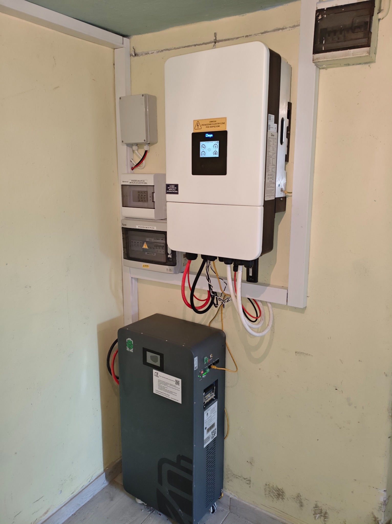 Kompletna instalacja: inwerter z wyświetlaczem, rozdzielnice AC/DC i akumulator energii na kółkach, zamontowane na ścianie w pomieszczeniu. Widoczne przewody i złącza.