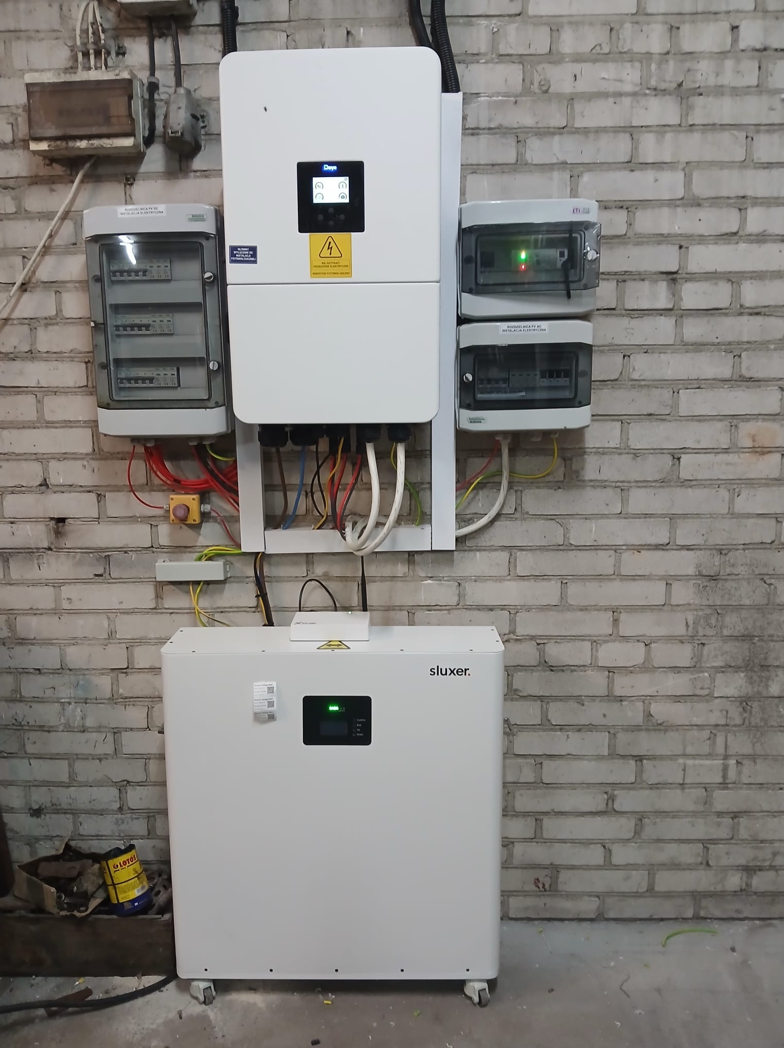 Zestaw urządzeń do instalacji OZE na tle ceglanej ściany: inwerter, akumulator energii Sluxer, rozdzielnice elektryczne. Widoczne okablowanie i elementy montażowe.