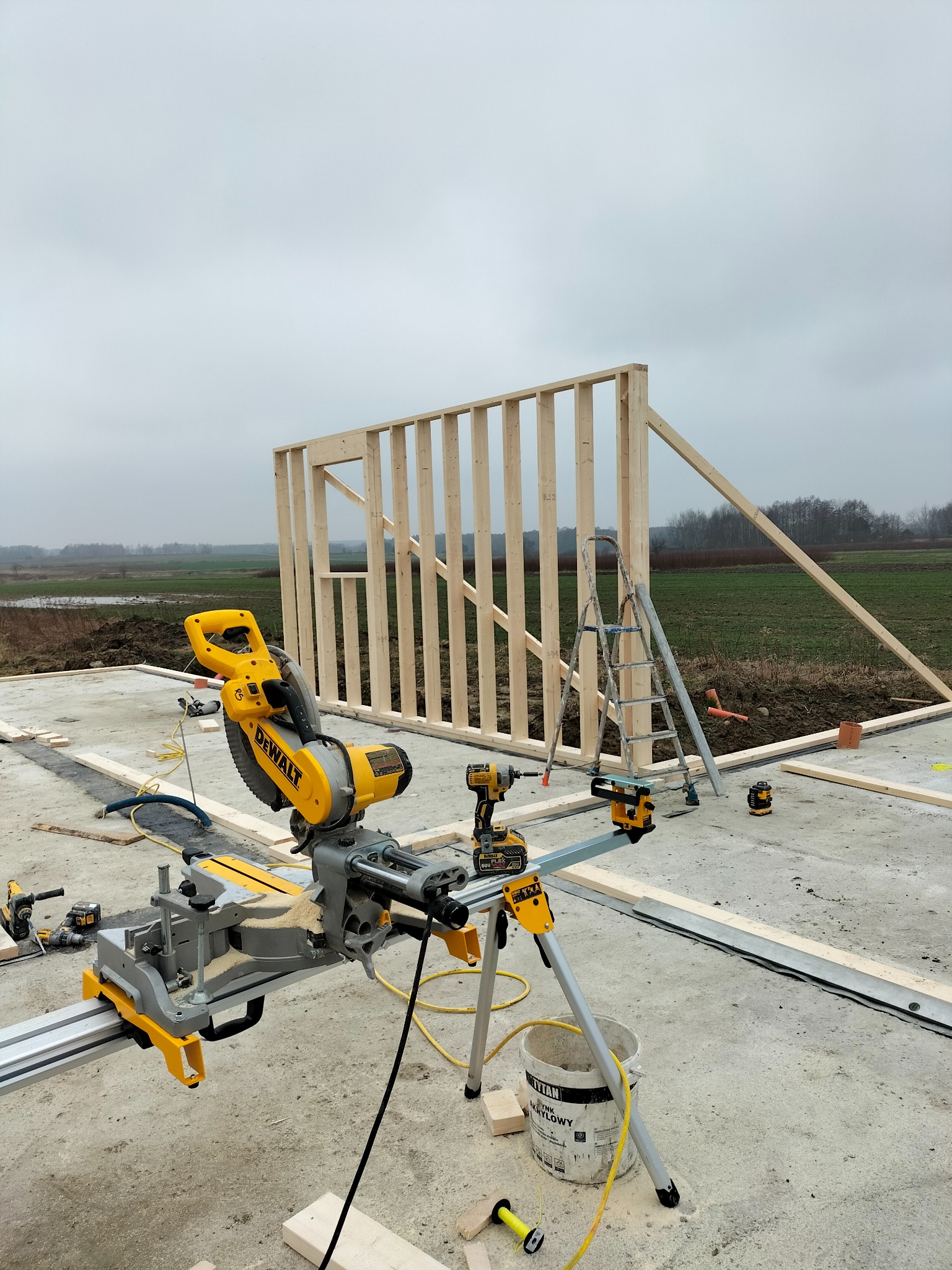 Wznoszenie drewnianej konstrukcji szkieletowej z użyciem profesjonalnych narzędzi DeWalt. Widoczny fragment ściany z oknem, piła ukośnica i wiertarko-wkrętarka.